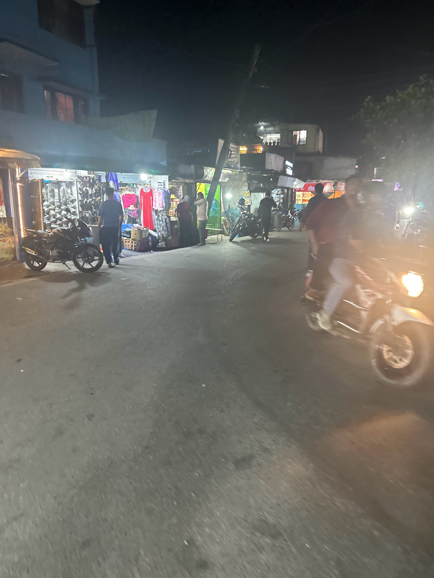 The image captures a nighttime street scene in Lalitpur, Nepal, with a focus on a roadside market and the flow of traffic. In the foreground, a motorcycle with its headlights on is prominent, partially blurred due to motion. The road surface is asphalt, appearing worn.
To the left, a street stall is visible, displaying an assortment of items, possibly clothing or accessories, arranged on shelves. A dark-colored motorcycle is parked in front of this stall. Further down the street, another stall features racks of clothing, including red dresses. Several people are present near the stalls, some appearing to be vendors and others customers. One man in a dark blue shirt and pants walks away from the camera.
In the background, buildings with lit windows suggest residential or commercial activity. A utility pole with wires is visible, contributing to the urban landscape. The overall lighting is dim, characteristic of nighttime, with artificial lights from the stalls and vehicles creating illuminated areas. The atmosphere suggests a bustling local market operating after dark.