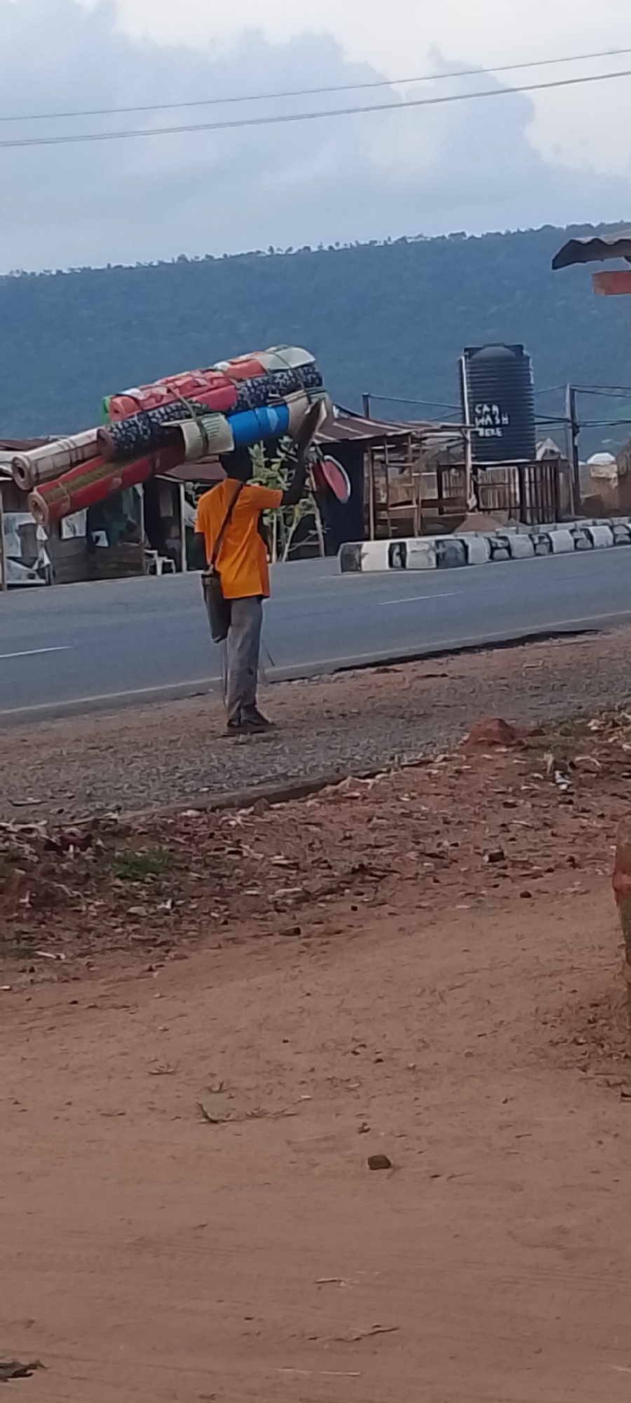 In Adankolo, Nigeria, a man stands on a gravel shoulder beside a paved road. He is carrying a large bundle of rolled-up mats or rugs on his head. The bundle is multicolored. The man is wearing an orange shirt, gray trousers, and sandals. A shoulder bag hangs from his left shoulder. Behind him, across the road, is a cluster of roadside structures, including a car wash with a large black water tank. A mountain range is visible in the background. The ground in the foreground is dirt and gravel.