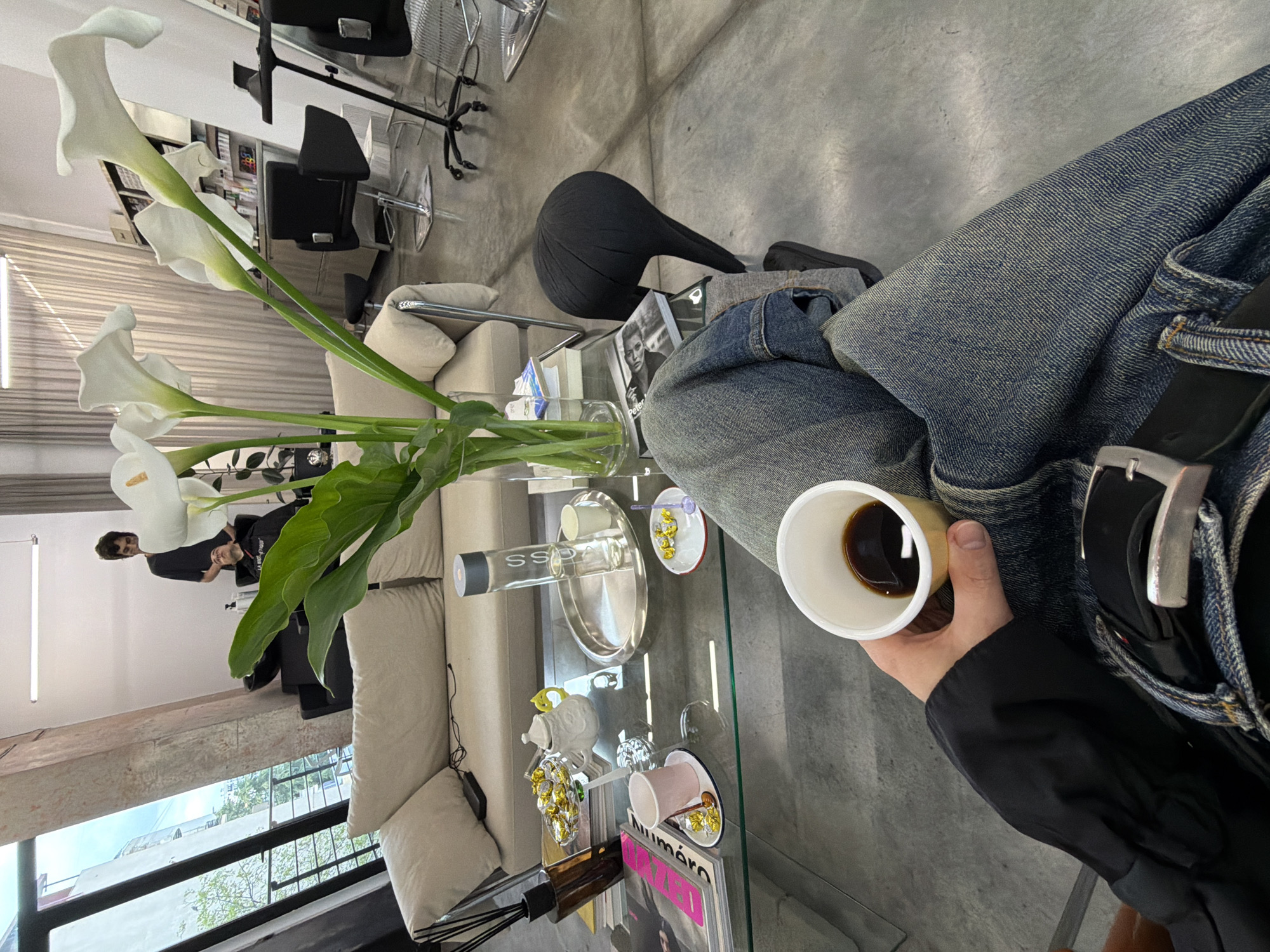 The image captures an interior scene, likely a modern lounge or waiting area, possibly within a salon or studio in Tbilisi, Georgia. The perspective is from a seated individual, looking down at their lap and a glass coffee table.
In the immediate foreground, a hand holds a white and light yellow paper cup containing dark coffee. The person is wearing faded blue jeans with a black belt and a silver buckle, and a black long-sleeved garment.
A clear glass coffee table dominates the center of the frame. On it, a tall glass vase holds a striking bouquet of white calla lilies with long green stems. Other items include a tall glass bottle bearing the label "SSPA," a shiny silver oval tray, small decorative dishes (one filled with yellow, jewel-like candies), a dark-colored diffuser with reeds, and two magazines. One magazine clearly displays "numéro" on its cover, while another nearby has "Peter Lindbergh" partially visible.
In the mid-ground, a light beige sofa with matching cushions provides seating. Behind it, a person, likely male, is seated, facing towards the right. Further back, several black, modern salon chairs with chrome bases are arranged, suggesting the establishment's function. The floor is a uniform light grey, possibly concrete. A long, rectangular fluorescent light fixture illuminates the background wall. Through a large window on the left, green trees and parts of an exterior building facade are visible, indicating a daytime setting in an urban environment. The overall atmosphere appears calm and contemporary.