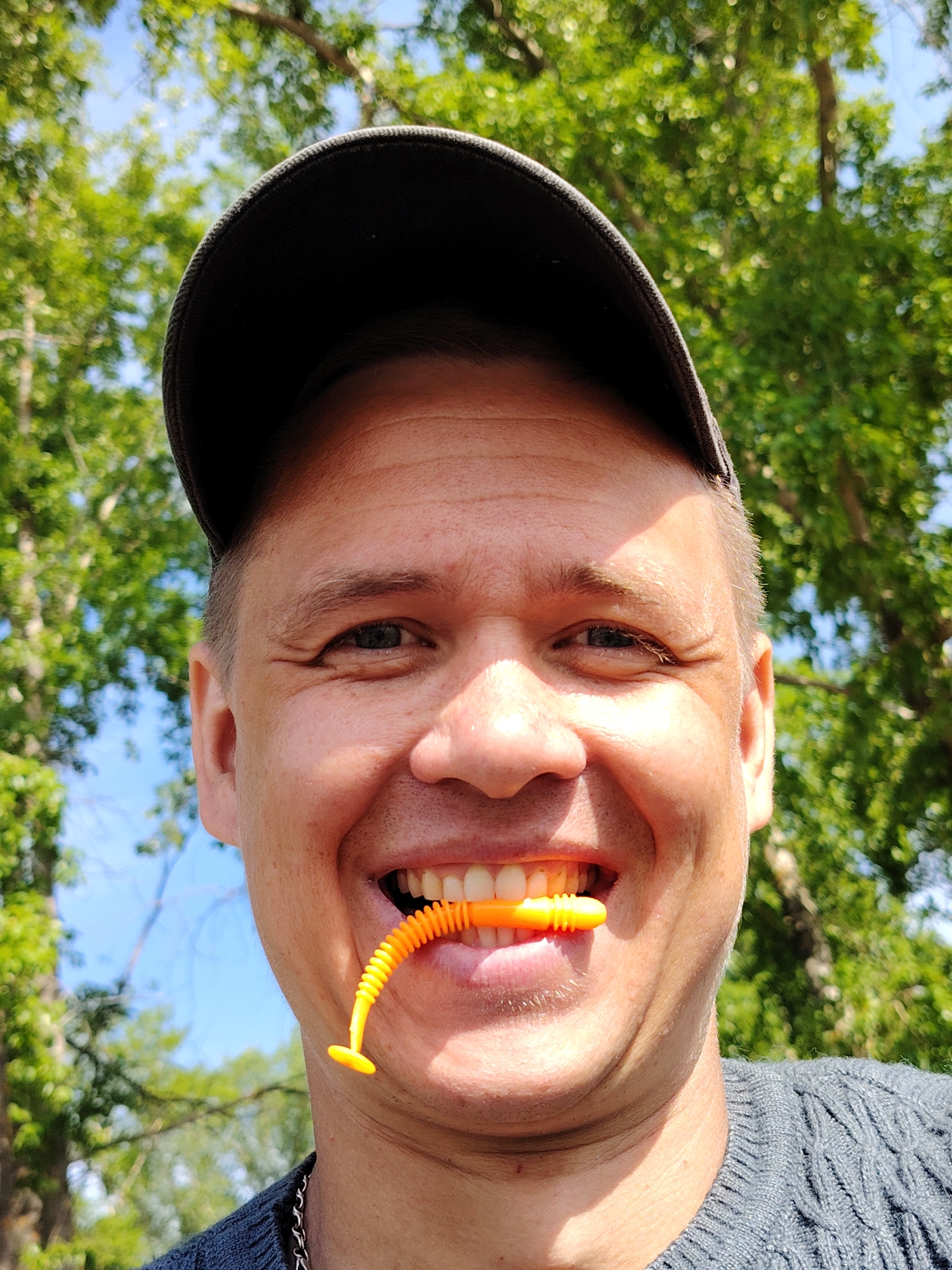A medium-close outdoor shot depicts a smiling man holding an orange object in his mouth. He is wearing a dark-colored baseball cap and a grey knit sweater with a chain. The background shows green trees and a blue sky. The man appears to be enjoying the outdoors, as indicated by his smile. There is no information to ascertain the location of this image, or the city of Chaikovsky.