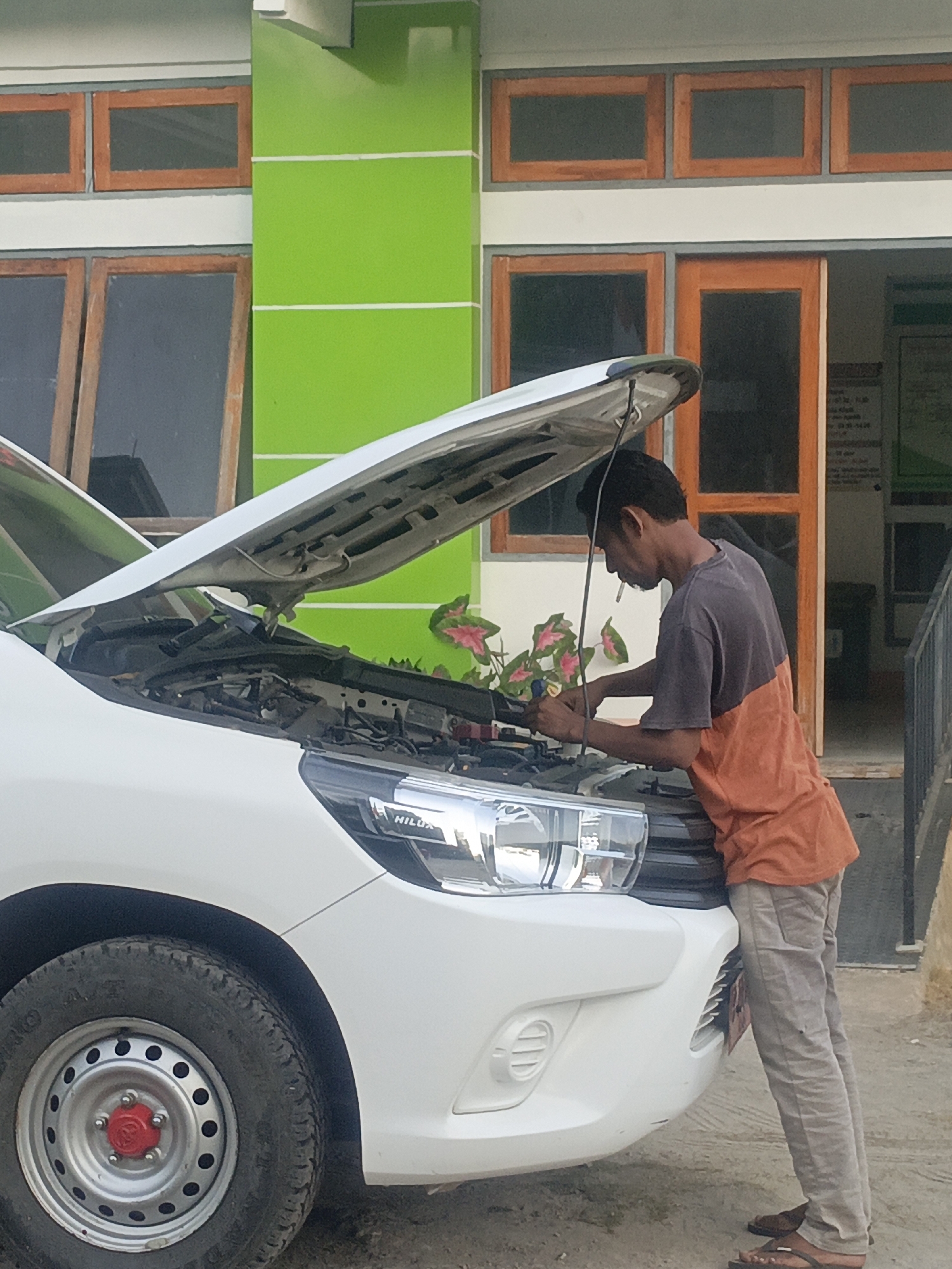 In the city of Solor Barat, Indonesia, a man is working on the engine of a white vehicle with its hood open. He is wearing a two-toned shirt, grey on the top and orange on the bottom, and light-colored pants. He appears to be holding a tool in his right hand and has a cigarette in his mouth. The vehicle is parked in front of a building with a vibrant green facade and wooden-framed windows. Some potted plants with pink and green leaves are visible near the car. The ground is dirt and sand. A street sign is partially visible in the background.
