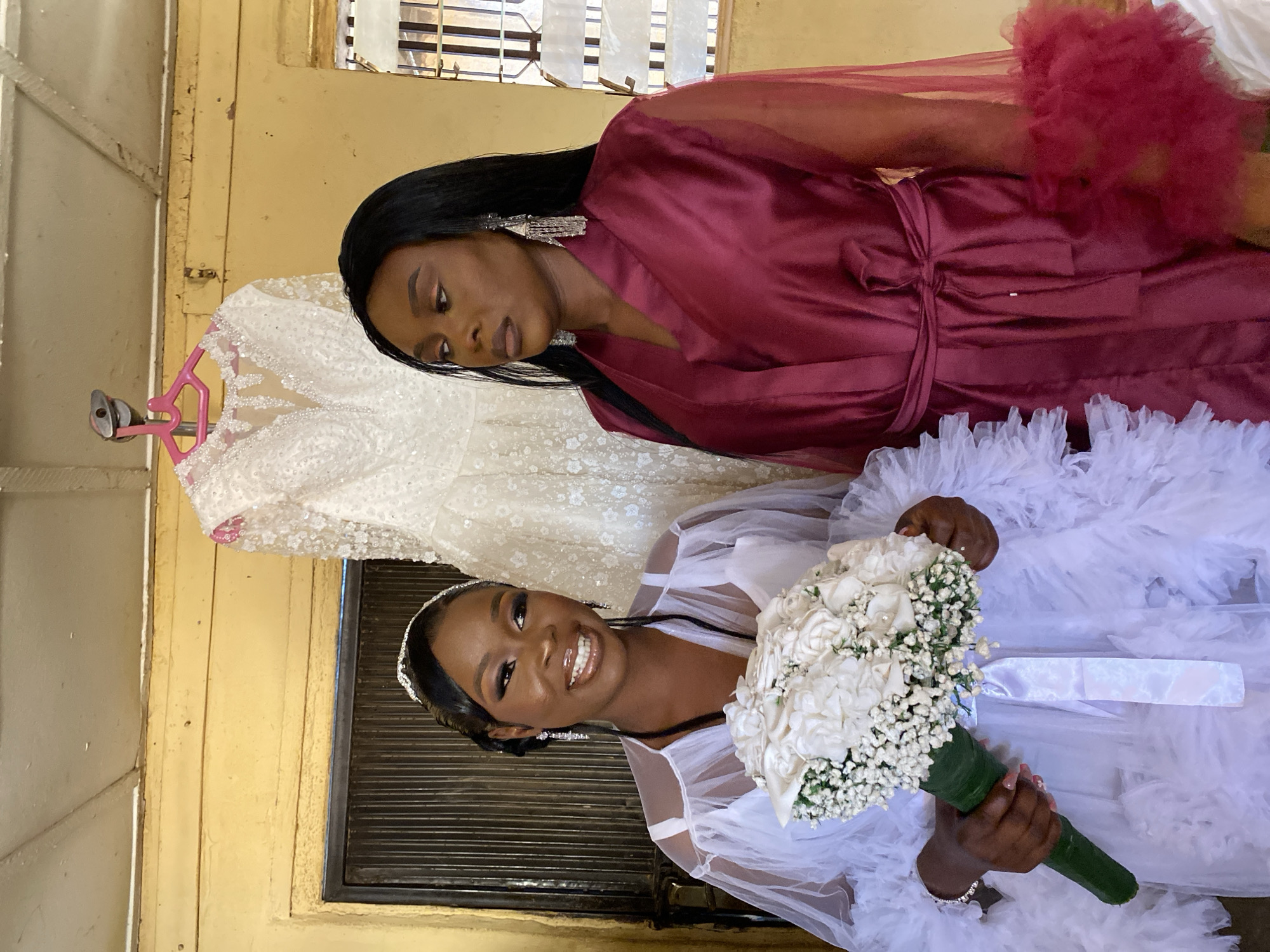 Two women pose for a photo indoors in Vom, Nigeria. One woman in a white, ruffled robe and a tiara holds a bouquet of white flowers. She smiles at the camera. The other woman, wearing a burgundy robe with furry red trim, looks down at the first woman. Behind them hangs a white wedding dress on a pink hanger. The background includes a window with blinds, a door, and a yellow wall.