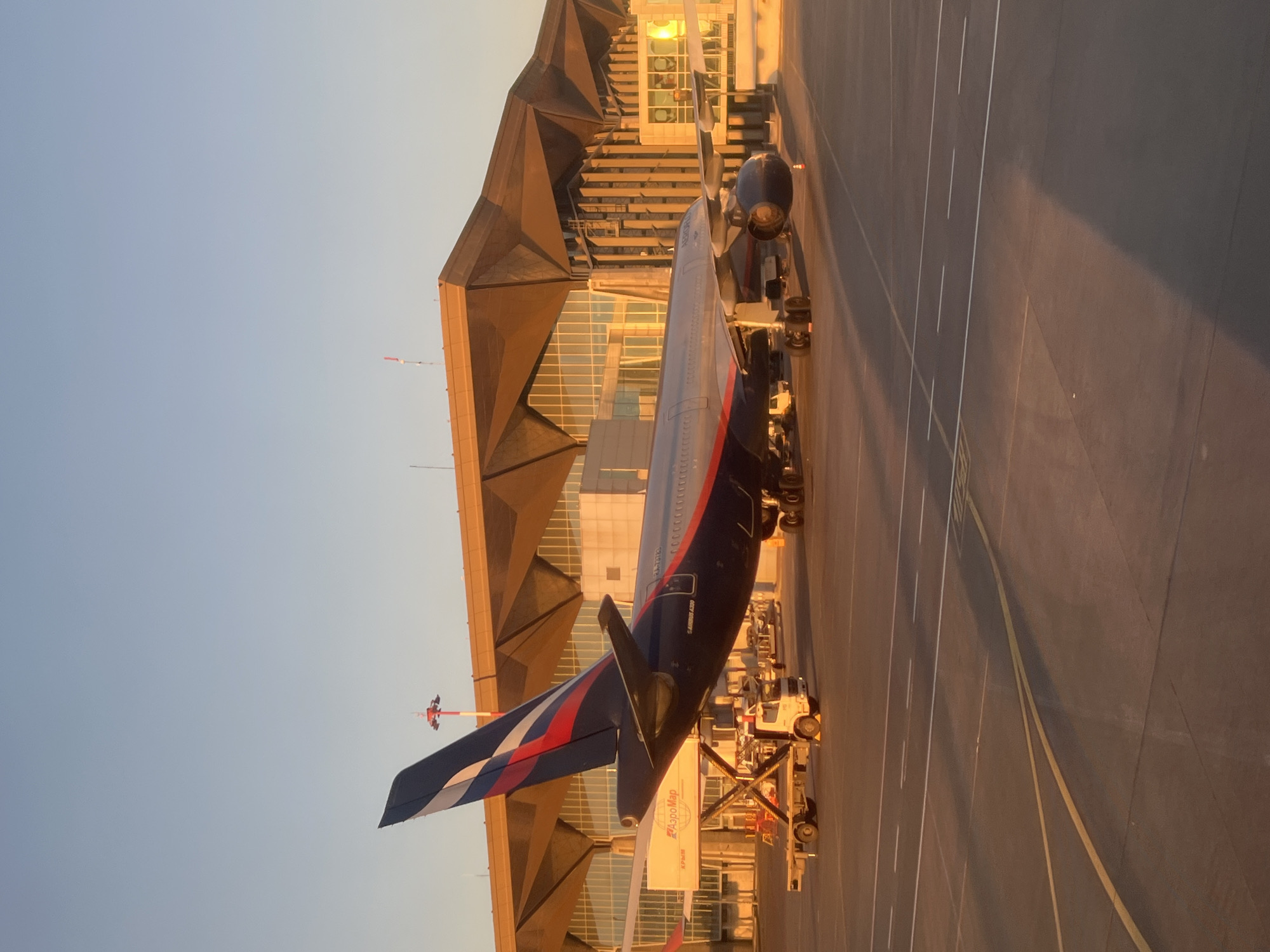 The image shows an Aeroflot Airbus A320 aircraft parked at Pulkovo Airport (LED) in Saint Petersburg, Russia. The plane is connected to a jet bridge, and airport ground service vehicles surround it. The airport terminal building is visible in the background. The sky is clear and light, indicating either dawn or dusk. The scene appears calm and orderly, likely depicting the process of boarding or disembarking passengers.