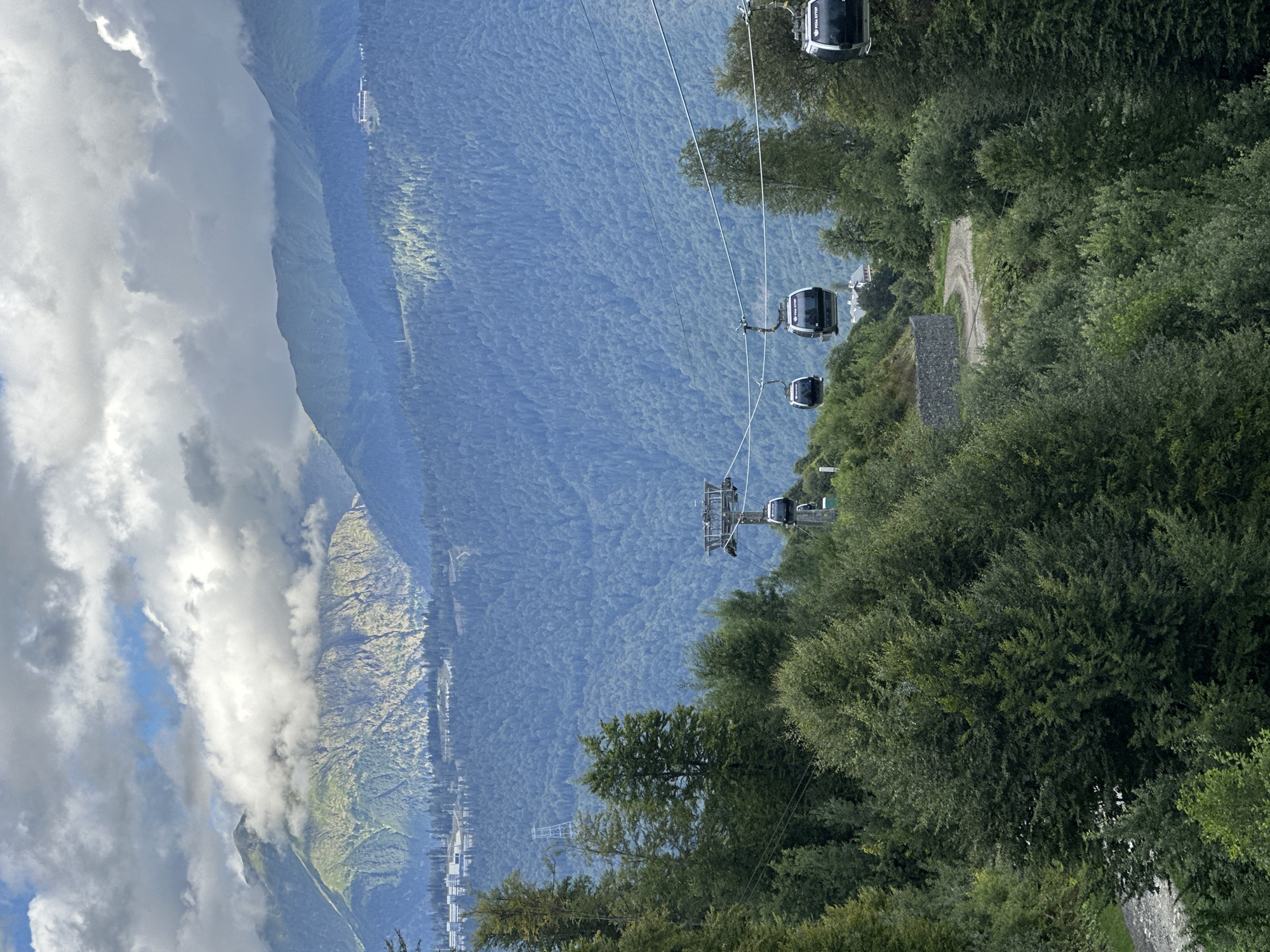 In Estosadok, Russia, a scenic view of the Caucasus Mountains unfolds under a sky filled with dramatic clouds. A cable car system traverses the landscape, with several gondolas suspended above a dense, verdant forest that blankets the lower slopes. The cable cars, dark and modern, are connected by thick cables stretching across the frame. In the distance, higher mountain peaks rise, some shrouded in mist and clouds, while others are illuminated by patches of sunlight. A winding dirt road is visible amidst the trees, and a few buildings can be seen nestled in the mountainside. The overall scene captures the grandeur of the natural landscape and the presence of human infrastructure within it.
