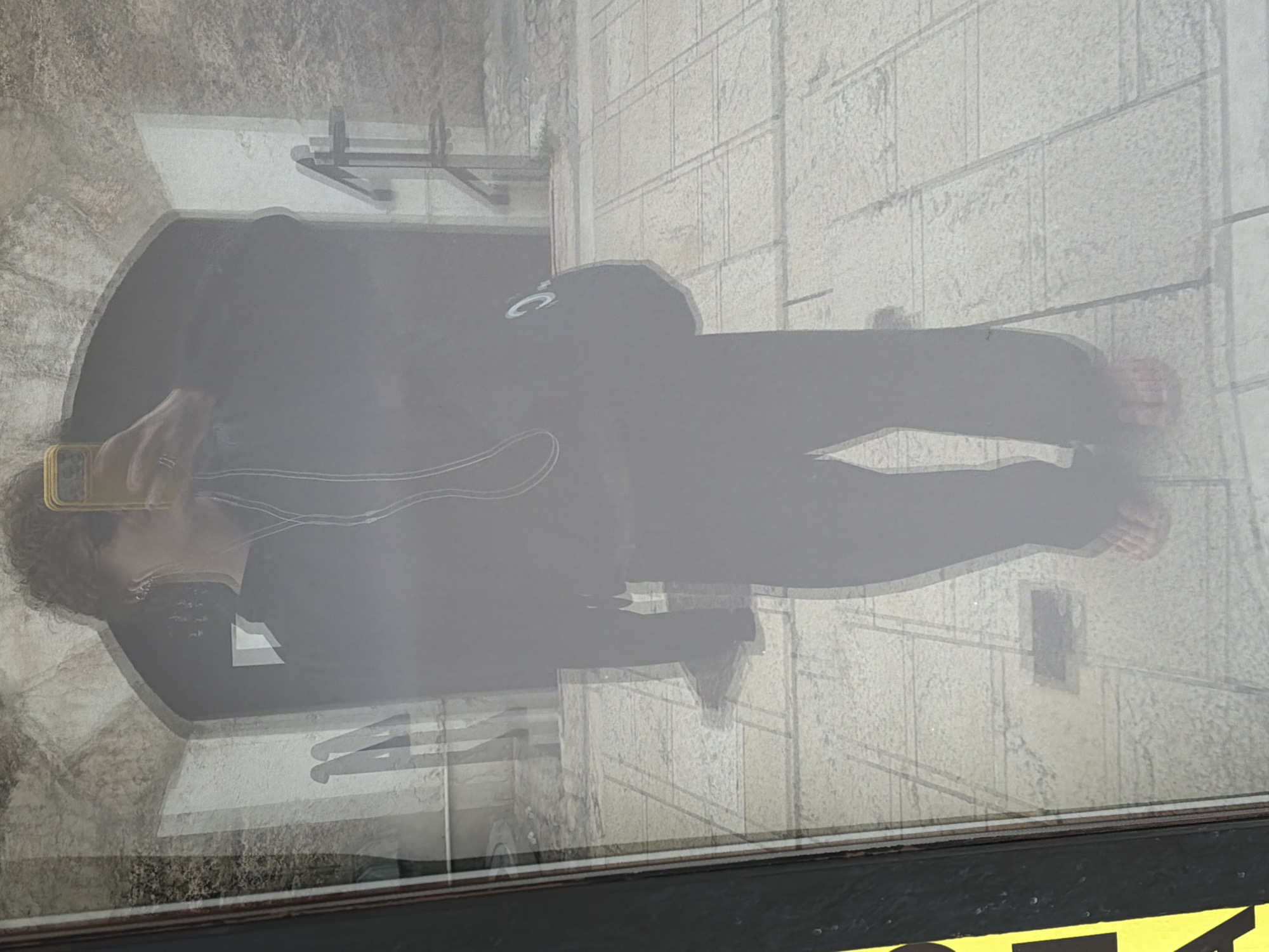 The image shows a reflection of a person holding a yellow smartphone in front of their face, taking a selfie. The person is wearing dark clothing, possibly a hoodie and pants, and has curly hair. They are also wearing white earphones. The reflection shows them standing on a paved area with square tiles. In the background of the reflection, there appears to be an arched doorway and some stonework, suggesting an old building or historical site. The overall image has a slightly hazy or distorted quality, common in reflections. The question specifies the location as Sarajevo, Bosnia and Herzegovina, though the visual elements alone do not provide definitive proof of this location without additional context. The presence of a yellow sign with black lettering on the left side of the image suggests it might be a street scene or a building exterior.