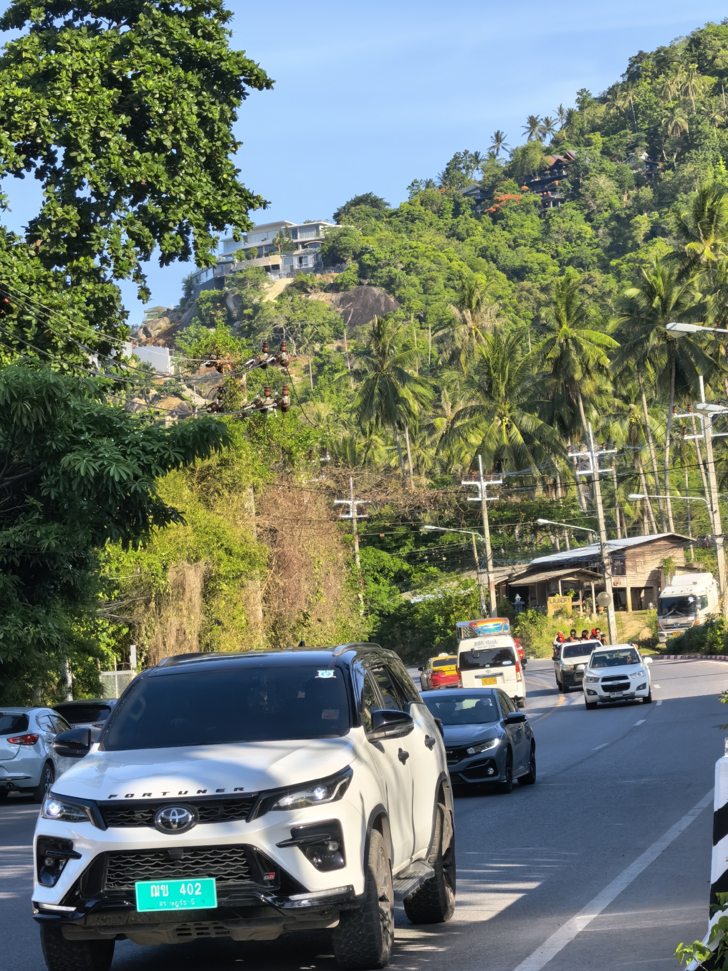 This is a vertical, eye-level, outdoor, daytime shot of a road in Ko Samui District, Thailand, on a clear day with a bright blue sky. The road is paved and has lane markings. On the left side of the road, a white Toyota Fortuner is prominently featured, facing forward. Its license plate is visible and reads "ญข 402". Behind the Fortuner, other vehicles are visible, including a white sedan and a dark-colored car.
The road curves to the right, leading uphill towards a lush, green hillside covered in dense vegetation and palm trees. Several houses and buildings are nestled into the hillside, with modern, multi-story structures visible at higher elevations and more rustic wooden buildings closer to the road. Power lines and utility poles run along the side of the road. In the middle ground, a white van with a colorful decal on top is stopped, with a yellow taxi behind it. A white pickup truck is also visible further up the road. The scene depicts a busy street with traffic on a sunny day in a tropical environment.