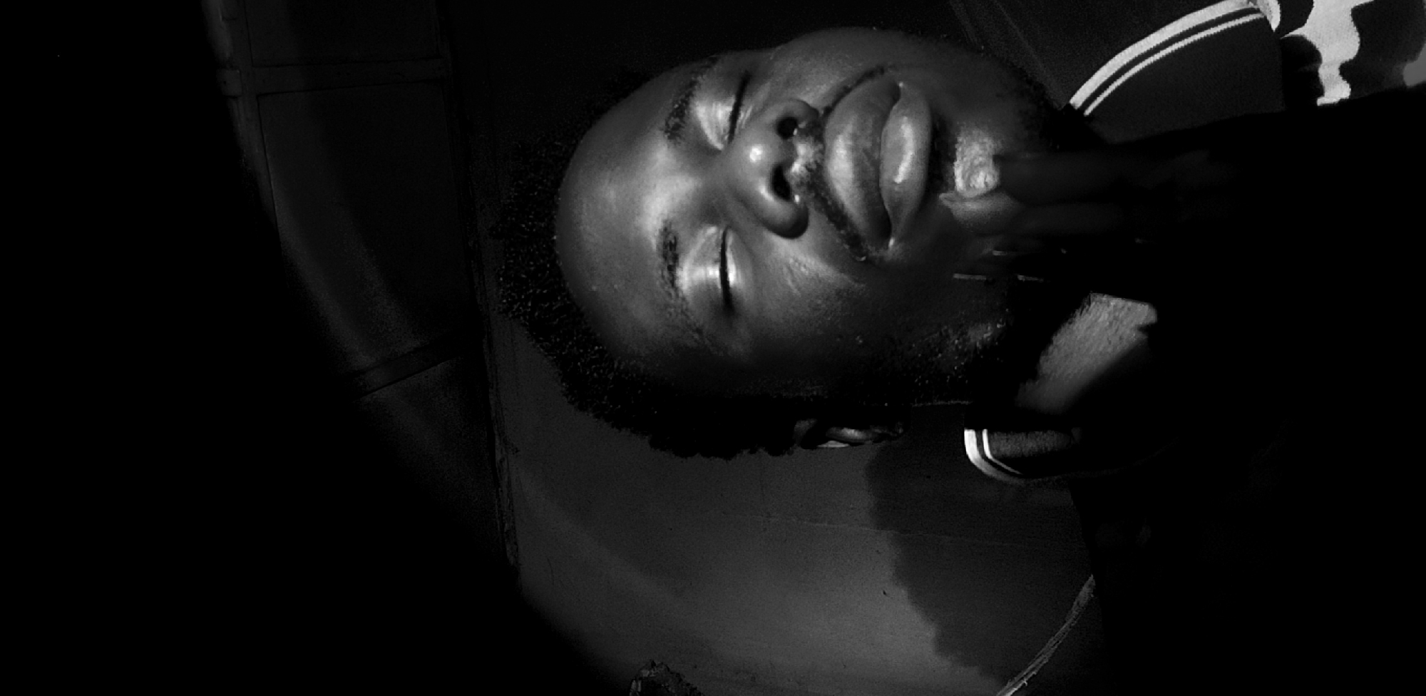 The image, taken in Narayi, Nigeria, is a black and white close-up of a young man with his eyes closed. He appears to be deep in thought or prayer, with his chin resting on his hand. Strong lighting casts dramatic shadows across his face and the surrounding dark environment, highlighting the contours of his features. He is wearing a collared shirt with striped detailing on the collar and sleeves. The background is dark and indistinct, suggesting an indoor setting with limited light sources.