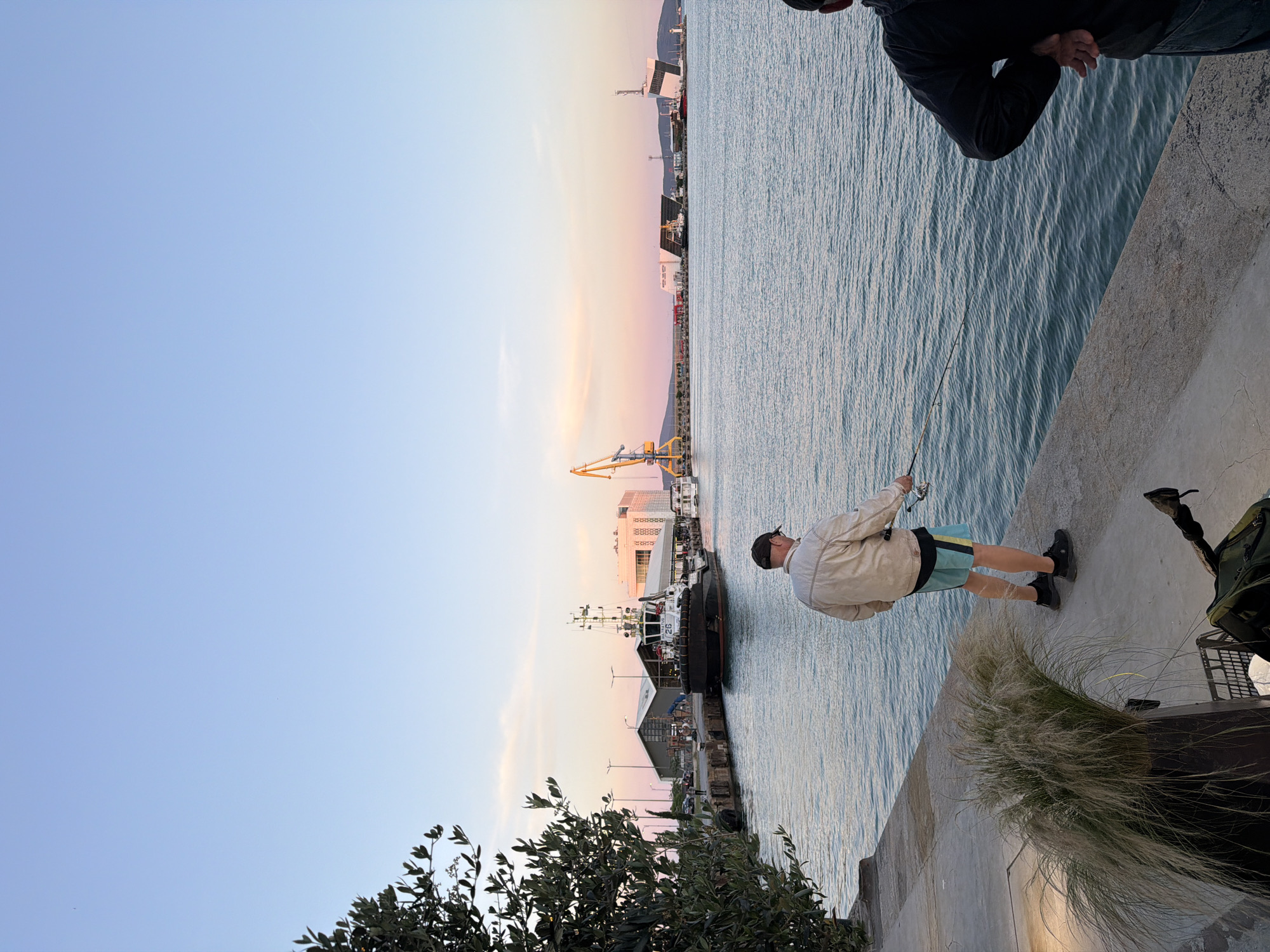 The image depicts a scene at the port of Burgas, Bulgaria, during what appears to be sunset. The sky displays a gradient of pale blue to soft pink hues. In the foreground, two individuals are visible. The person on the left is facing away from the camera, standing on a concrete embankment and fishing with a rod and reel. This individual is wearing a light-colored jacket, blue and yellow shorts, and dark shoes, along with a baseball cap. To the right of the fisherman, another person, also facing away, is partially visible, wearing a dark jacket and blue jeans. A prominent bushy plant in a dark container sits on the embankment to the left, and fishing gear is placed on the ground near it.
In the mid-ground, the calm water of the port is visible, with gentle ripples on its surface. A large tugboat, identified by the number '26' on its side and the text 'ULTRATUG', is docked alongside the embankment. Several other vessels and structures are present in the port. Further in the background, a yellow construction crane stands tall, and various industrial buildings and a white modern building with a distinct geometric design are situated along the waterfront. The distant horizon shows a range of low hills or mountains. The overall atmosphere is tranquil, suggesting a quiet evening at the port.