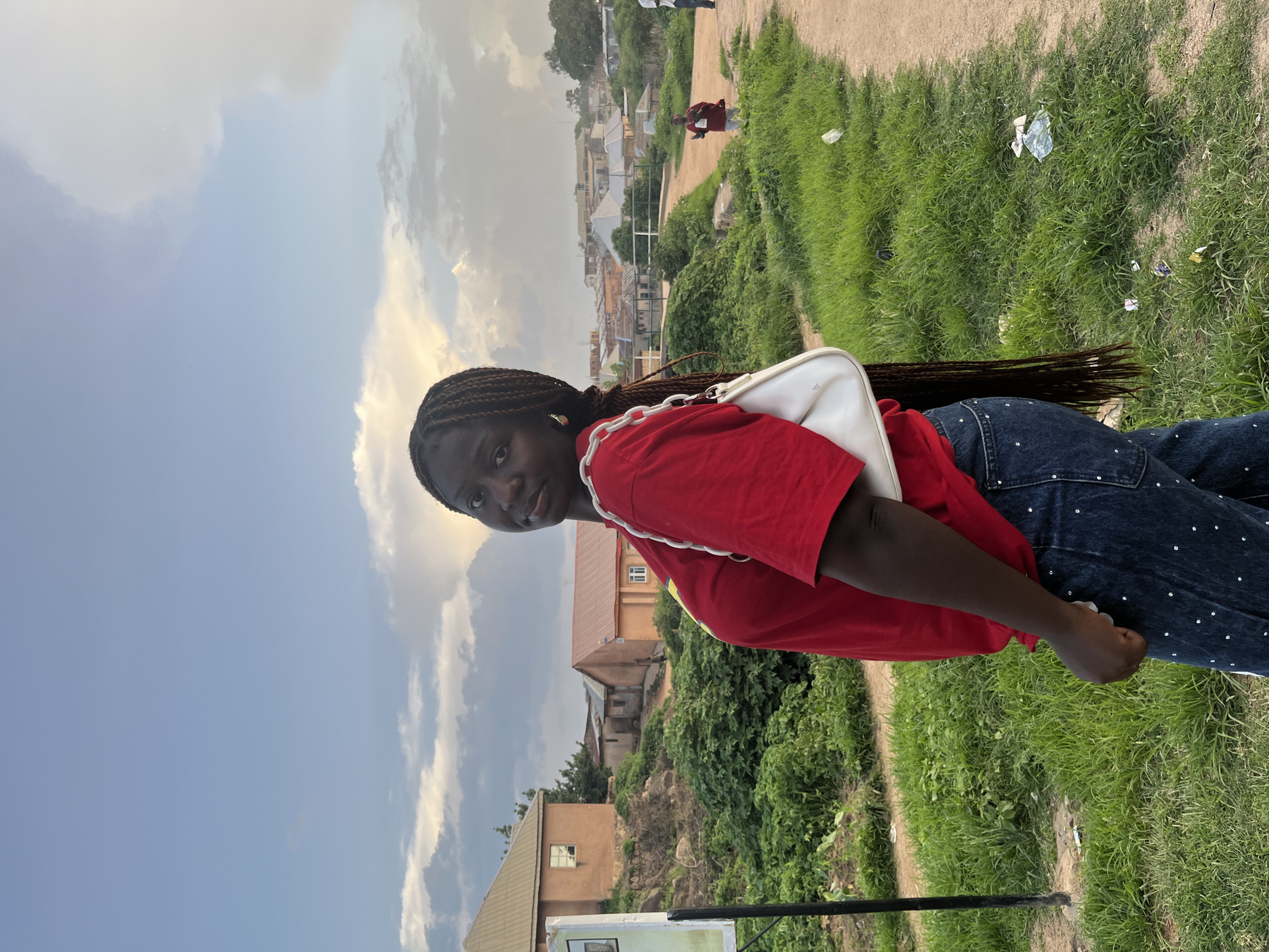A young Black woman with long, dark braided hair and wearing a red t-shirt and dark, bejeweled jeans stands on a dirt path in a residential area of Jos, Nigeria. She is holding a white purse with a chain strap and looking over her left shoulder at the camera. The background shows several houses with various colored roofs, dense green vegetation, and a cloudy sky with hints of sunlight. The path is lined with green grass and some scattered litter.
