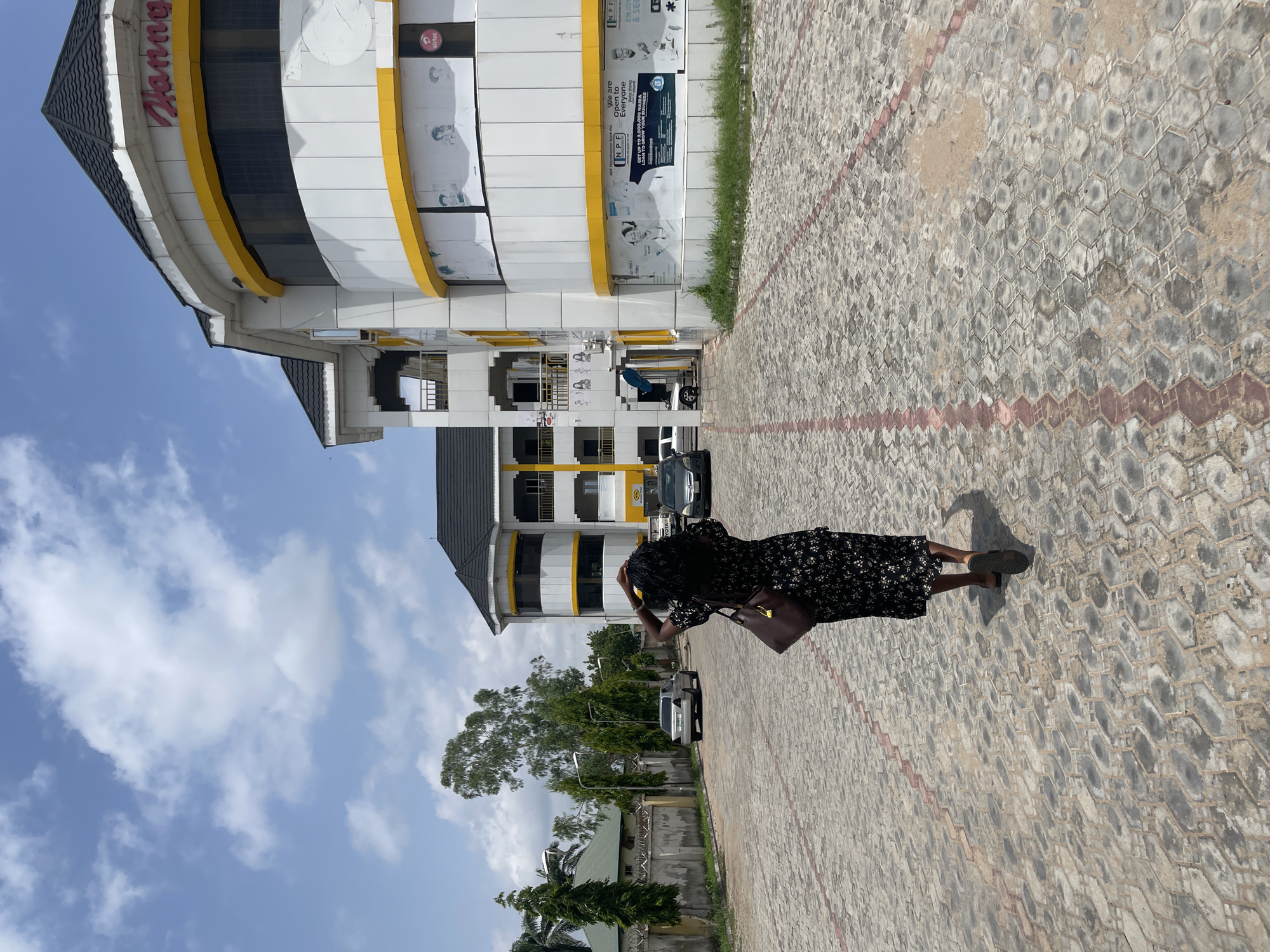 In Jalingo, Nigeria, a woman with dark, curly hair walks away from the camera on a paved outdoor area. She is wearing a dark, floral-patterned dress and carrying a brown shoulder bag. The ground is covered in interlocking hexagonal stones with red and pink markings. To the right, a modern, multi-story building with white and yellow accents is visible, featuring the name "Hanna" in red lettering at the top. Various posters and advertisements are affixed to the building's facade. Several cars are parked in the background, and trees and other vegetation are scattered throughout the scene. The sky above is blue with scattered white clouds.