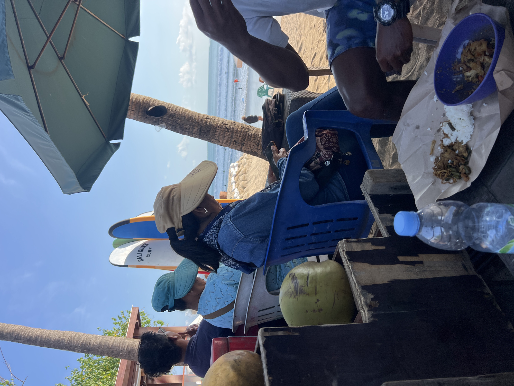 In Kuta, Indonesia, on a sandy beach under a bright blue sky, a group of people are seated in chairs, with surfboards visible in the background. The scene suggests a casual setting, possibly a beachfront eatery or relaxation area.
In the foreground, several objects are placed on a wooden surface. A clear plastic bottle of water with a blue cap stands upright. To the left, two coconuts are visible, one larger and lighter green, the other smaller and darker. A meal is spread out on brown paper, with white rice and a mixture of food items in a purple bowl and on the paper.
Several people are partially visible, seated in chairs. One person on the far left wears a dark shirt and has dark, curly hair. Next to them, a person in a turquoise hat is seated in a white chair, wearing a light blue patterned top. Another person, wearing a tan hat and a denim jacket, is in a blue chair, facing away from the camera. To the right, a person's arm and upper body are visible, wearing a white t-shirt and patterned blue shorts, with a wristwatch on their left wrist.
Behind the seated individuals, several surfboards stand upright. One surfboard has the text "BALIGUNS SURF" printed on it in black letters. Palm trees are present, and a large green umbrella provides shade. In the middle ground, the ocean with small waves is visible, with a few people in the water. The beach is sandy, with some light-colored sandbag-like structures in the distance. The overall ambiance is relaxed and summery.