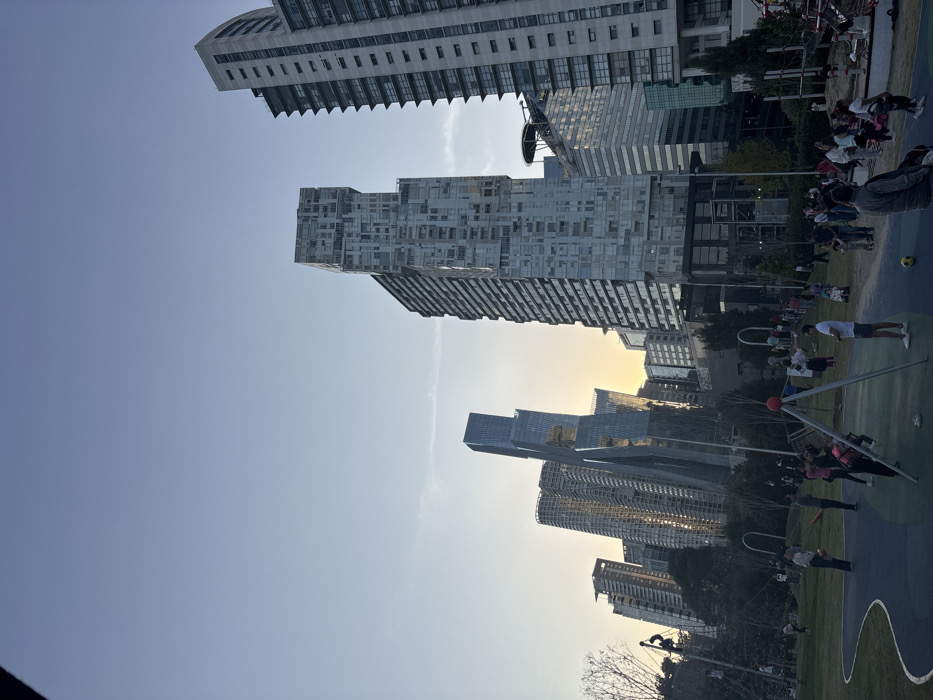 The image displays an urban landscape featuring a collection of modern high-rise buildings towering over a public park or recreational area. The original image is rotated 90 degrees clockwise, so for clarity, the description assumes a correct upright orientation where the buildings are vertical and the sky is at the top.
The sky occupies roughly the upper two-thirds of the frame, showcasing a clear, light blue expanse. Towards the right side, the sky transitions to a warm yellow and orange glow, indicating the low angle of the sun, suggestive of either late afternoon/sunset or early morning/sunrise. This warm light illuminates some of the buildings, casting soft hues on their facades.
Multiple distinctive high-rise buildings dominate the scene. On the left, a tall, light-colored building with numerous rectangular windows and a stepped or terraced design on its upper sections is prominent. In the central part of the skyline, another striking high-rise features a complex, fragmented facade composed of what appears to be irregularly stacked blocks or cubes made of grey and glass elements. Below this, a section with horizontal lines or balconies is visible. Further to the right, a cluster of sleek, modern buildings with extensive glass facades and varying heights catch the warm sunlight. Some of these structures exhibit unique architectural designs, including curved elements, contributing to a dense and contemporary urban skyline.
The lower third of the image depicts a public green space, likely a park or playground. The ground is predominantly green, interspersed with dark blue or black sections that could be pathways or specialized play surfaces. Several people are scattered throughout this area, engaging in various activities. In the foreground, towards the right, there's an outdoor exercise apparatus with a red spherical joint and silver bars, and a person wearing a dark top and light pants is actively using it. Another individual, dressed in a white t-shirt and dark shorts, stands nearby, seemingly holding a mobile phone. Further in the background of the park, a small group of people is visible, and a yellow ball (possibly a soccer ball) rests on the ground. Small trees and shrubs are also present, adding greenery to the urban environment, along with some dark metal structures that might be part of playground or additional exercise equipment.
The overall scene conveys a bustling urban setting where modern architecture meets public recreational spaces, under a clear sky with beautiful warm light from the low-lying sun. No discernible text is visible in the image.