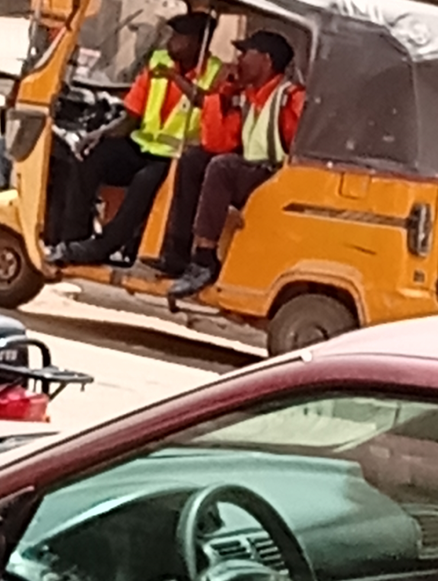 The image shows an auto rickshaw in what appears to be Maiburiji, Nigeria, occupied by two individuals wearing high-visibility vests. The rickshaw is predominantly yellow, and the occupants wear black caps and dark pants. The person on the left side is wearing a yellow vest with orange trim over an orange shirt. The other person is wearing a yellow vest with orange trim over an orange shirt. The rickshaw is positioned on a street, and a car with a maroon roof and a visible steering wheel is in the foreground, partially obscuring the view.