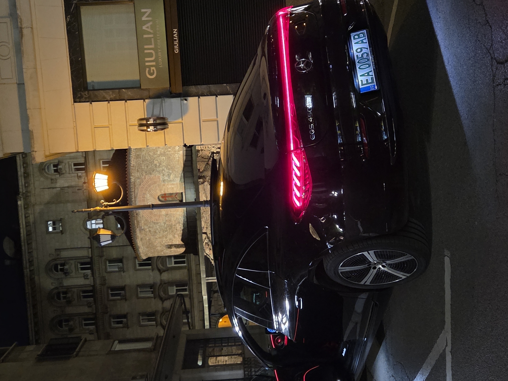 A dark Mercedes-Benz EQS 450 luxury sedan is prominently parked on what appears to be a city street or parking area at night. The car is angled, revealing its rear and left side, with its distinctive red LED taillights illuminated, casting a glow on the dark vehicle. The license plate, "EA 0059 AB" with the "BG" (Bulgaria) country code, is clearly visible and lit. The vehicle features multi-spoke alloy wheels.
In the background, two buildings with varying architectural styles are visible. One building, to the left of the car, has a more traditional European facade with arched windows and what looks like brickwork and decorative elements. Another building, slightly to the right and further back, has a modern storefront with a sign that reads "GIULIAN" and "LUXURY COLLECTION." A classic street light pole with two illuminated lamps stands between the car and the older building, providing additional light to the scene. The ground is asphalt with some faint white parking line markings visible. The overall impression is a vibrant urban night scene in Sofia, Bulgaria, as indicated by the license plate and provided context. No people are visible.