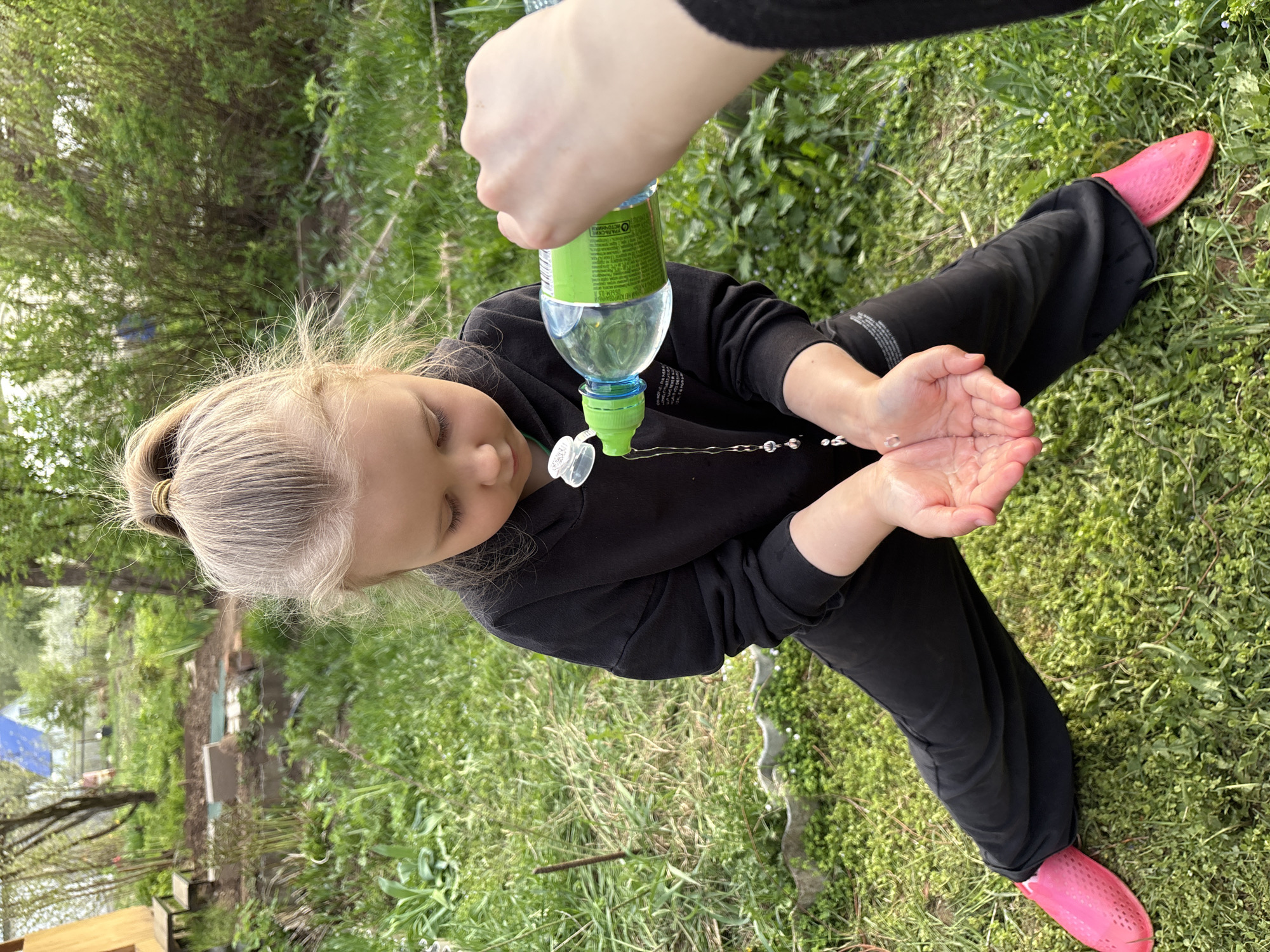 A young girl with blonde hair, wearing black pants and pink shoes, is standing on grass. An arm is pouring water from a plastic bottle into her outstretched hands. The background consists of green grass, plants, and trees. The photo appears to be taken outdoors, possibly in a garden or park. The location could be in Chaikovsky, Russia.