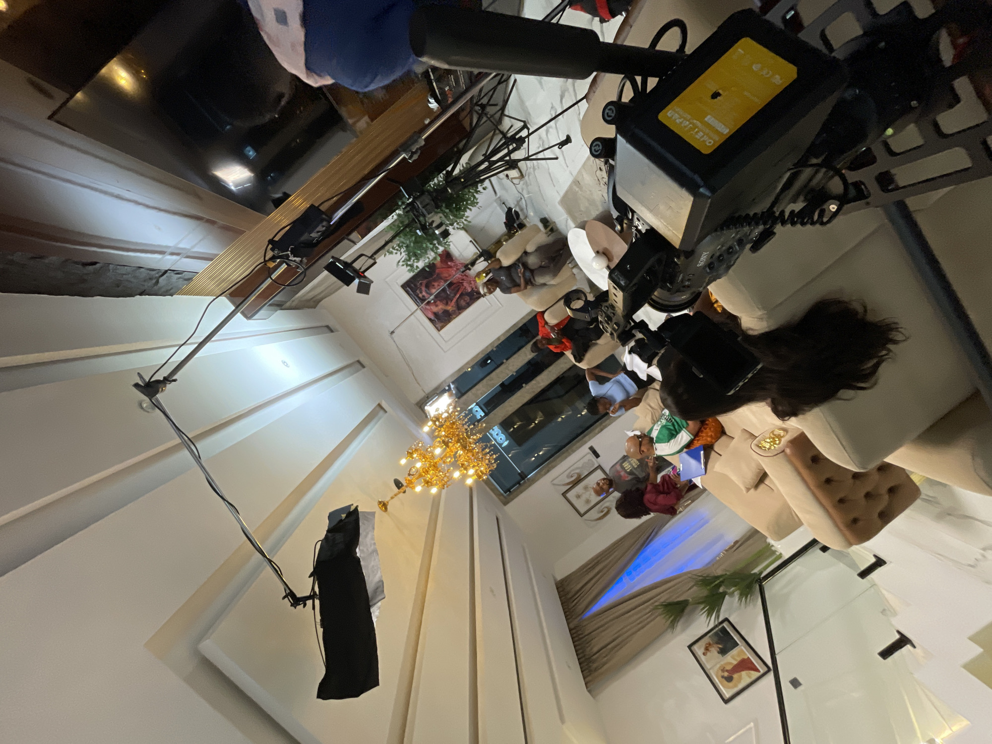 In an interior setting, a video production is underway. A boom mic extends from the ceiling, holding a softbox light. In the foreground, a video camera is mounted on a rig. Several people are seated on a large L-shaped sofa in the middle ground. One person, wearing a green and white jacket, is holding a blue folder. A chandelier hangs prominently from the ceiling, casting a warm glow. The room features modern architecture with recessed lighting and a large television screen visible in the background. Artwork is displayed on the walls. The scene suggests a casual interview or discussion taking place in a well-furnished living space.