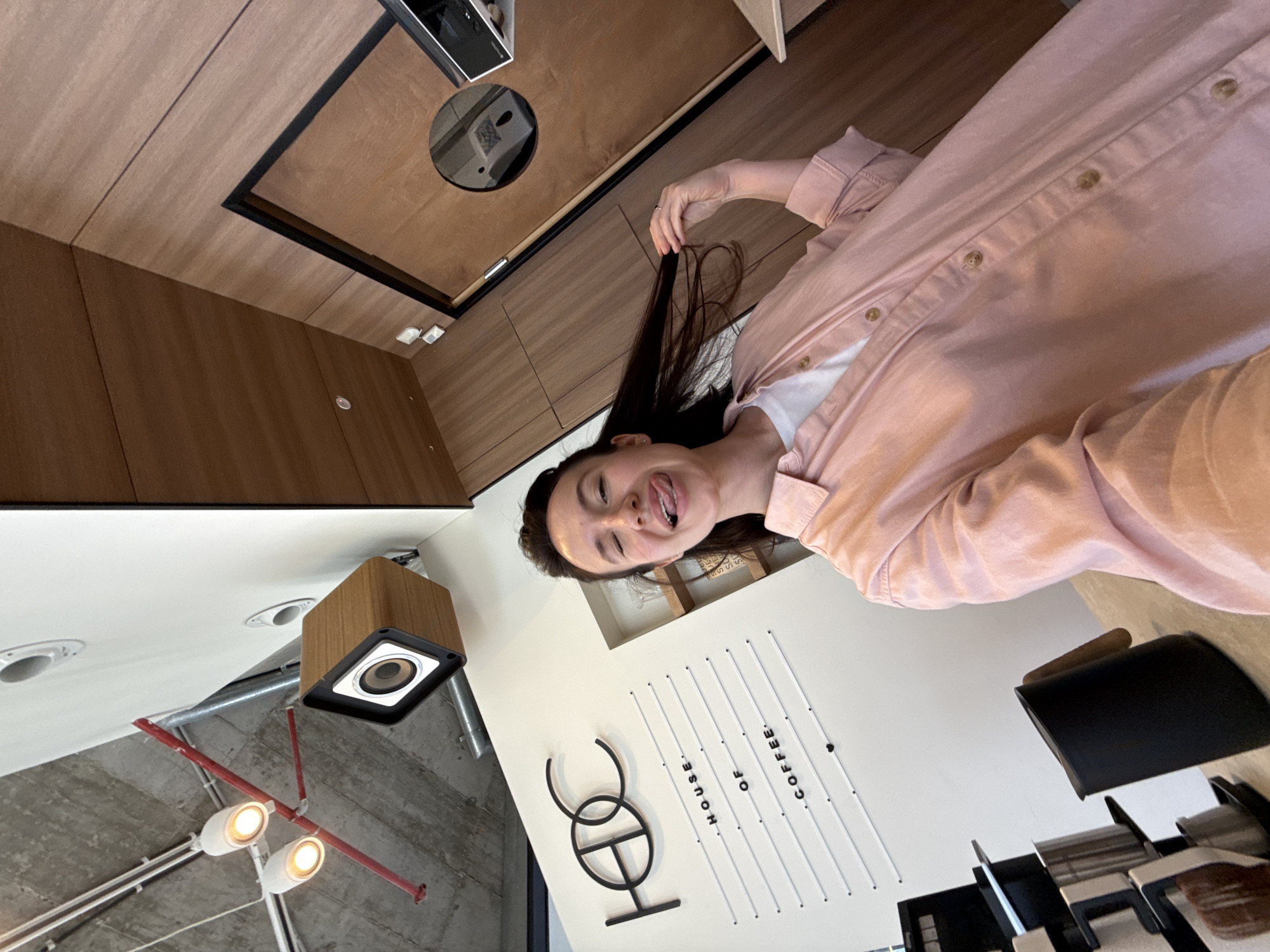 The image captures a playful moment with a young woman posing indoors, likely in a cafe in Tel-Aviv, Israel. She is seen from the chest up, looking directly at the camera with a wide smile and her tongue slightly extended. Her long, dark hair is held up by her left hand. She wears a light pink button-up shirt over a white t-shirt.
The setting is a modern establishment featuring a mix of warm wooden panels on the walls and ceiling, contrasted with a more industrial ceiling area revealing exposed concrete, red pipes, and track lighting with spotlights. Recessed lights are also visible in other ceiling sections. A wooden-encased speaker is mounted on a wall or ceiling.
A prominent feature in the background is a white wall displaying a sign. The text "HOUSE OF COFFEE" is spelled out vertically using small white pegs on a grid, and below it, a stylized black logo spells "HOC." In the lower right of the image, dark chairs and tables are visible, consistent with a cafe environment. A circular reflective surface or mirror is embedded in the wooden ceiling panel above the woman. The overall mood is casual and cheerful, suggested by the woman's playful expression.