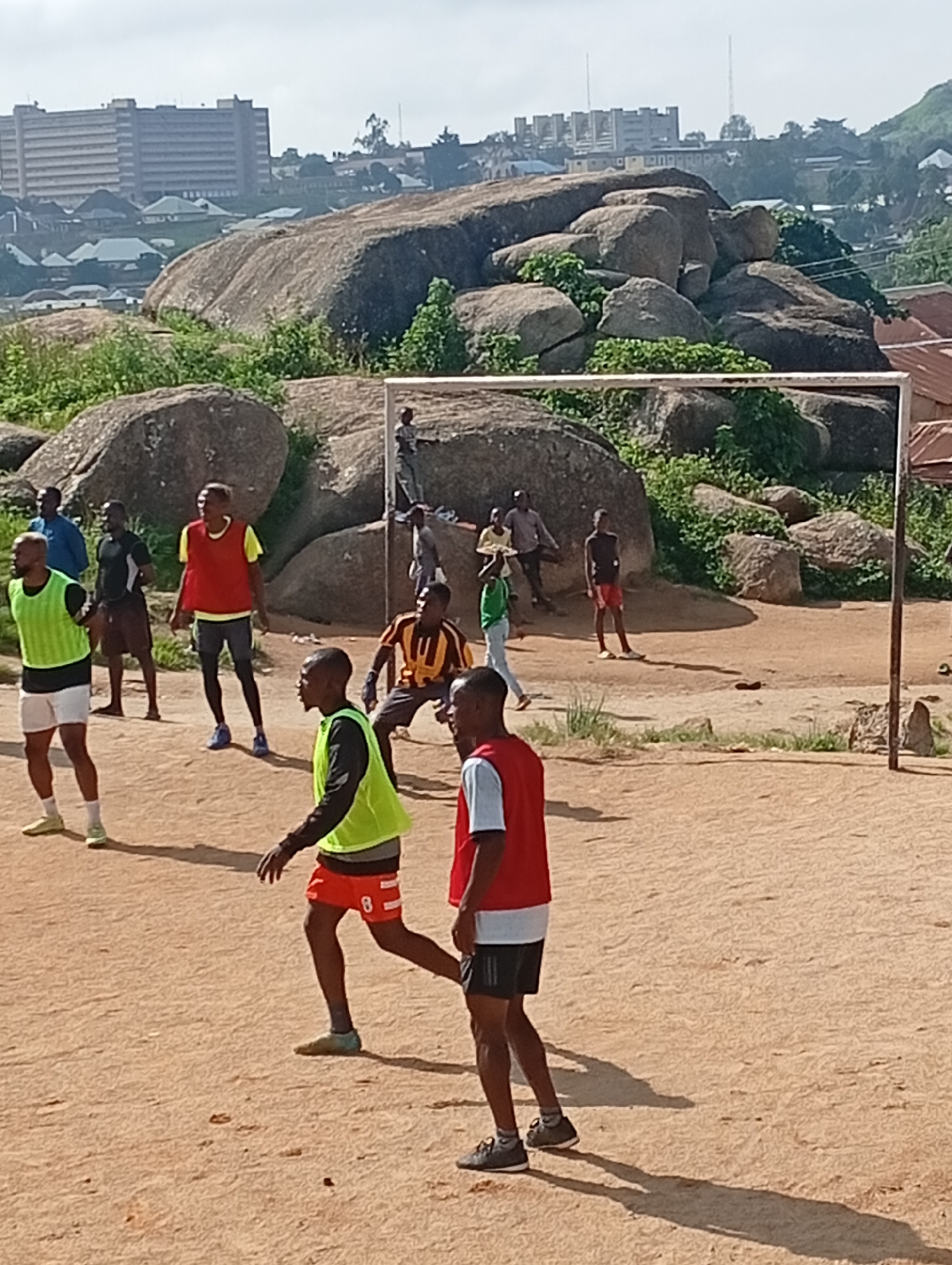In Jos, Nigeria, a group of young men are engaged in a soccer game on a dirt field situated amidst large rock formations. The players are wearing various athletic attire, including jerseys, shorts, and bibs. One player in the foreground wears a red bib over a white shirt and black shorts, while another in a green bib with black sleeves and orange shorts is in motion. In the background, a soccer goalpost stands erect. Beyond the rocky terrain, a cityscape with buildings and structures can be observed, suggesting an urban or semi-urban environment. The sky is overcast.