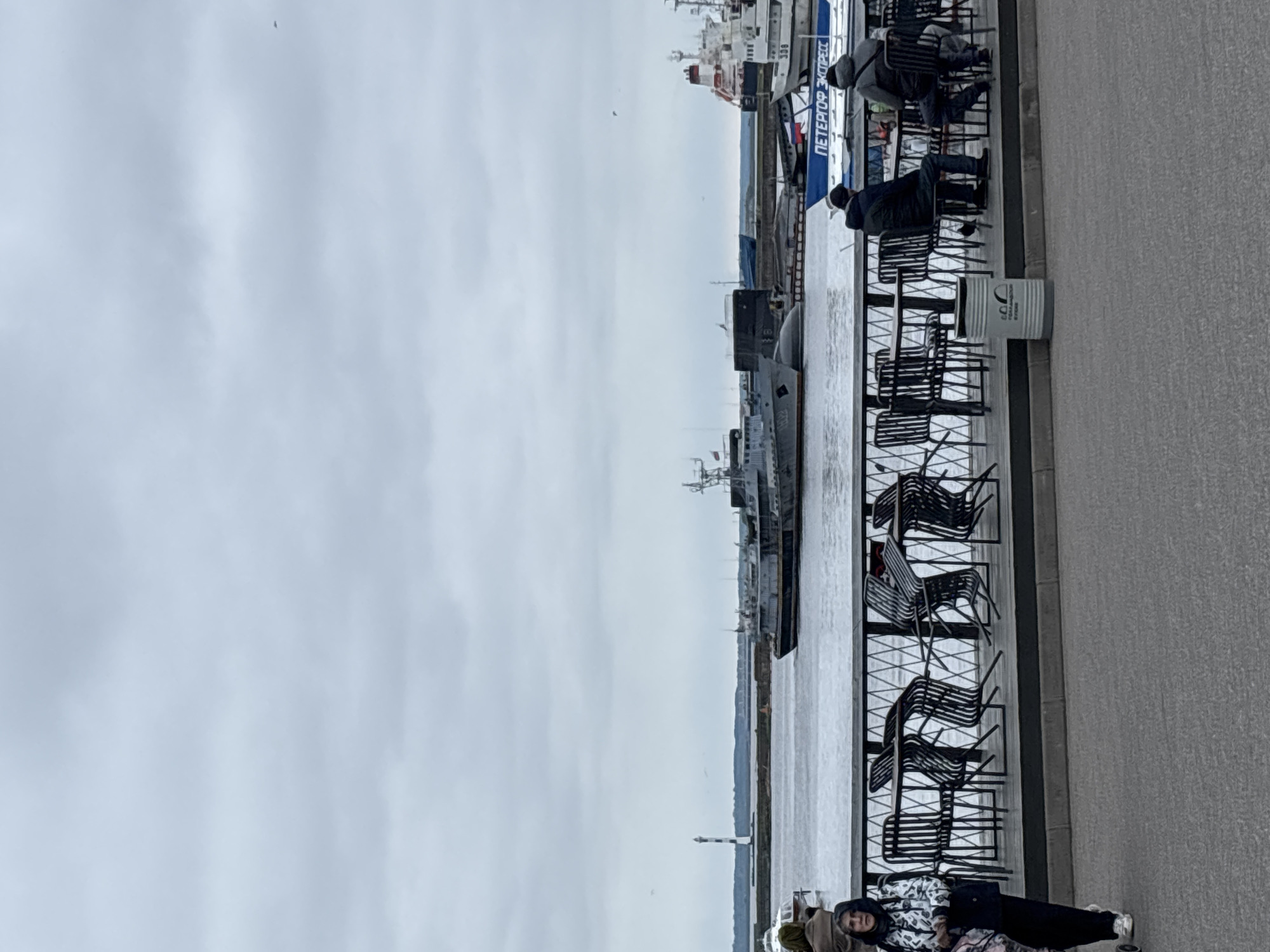 The image shows a waterfront scene in Kronstadt, Russia. A row of black metal chairs lines a grey pavement. People are seen sitting in the chairs, facing the water. Multiple ships and boats are docked at a pier along the waterfront. The sky is overcast and grey. A light house is visible on the left, across the water.