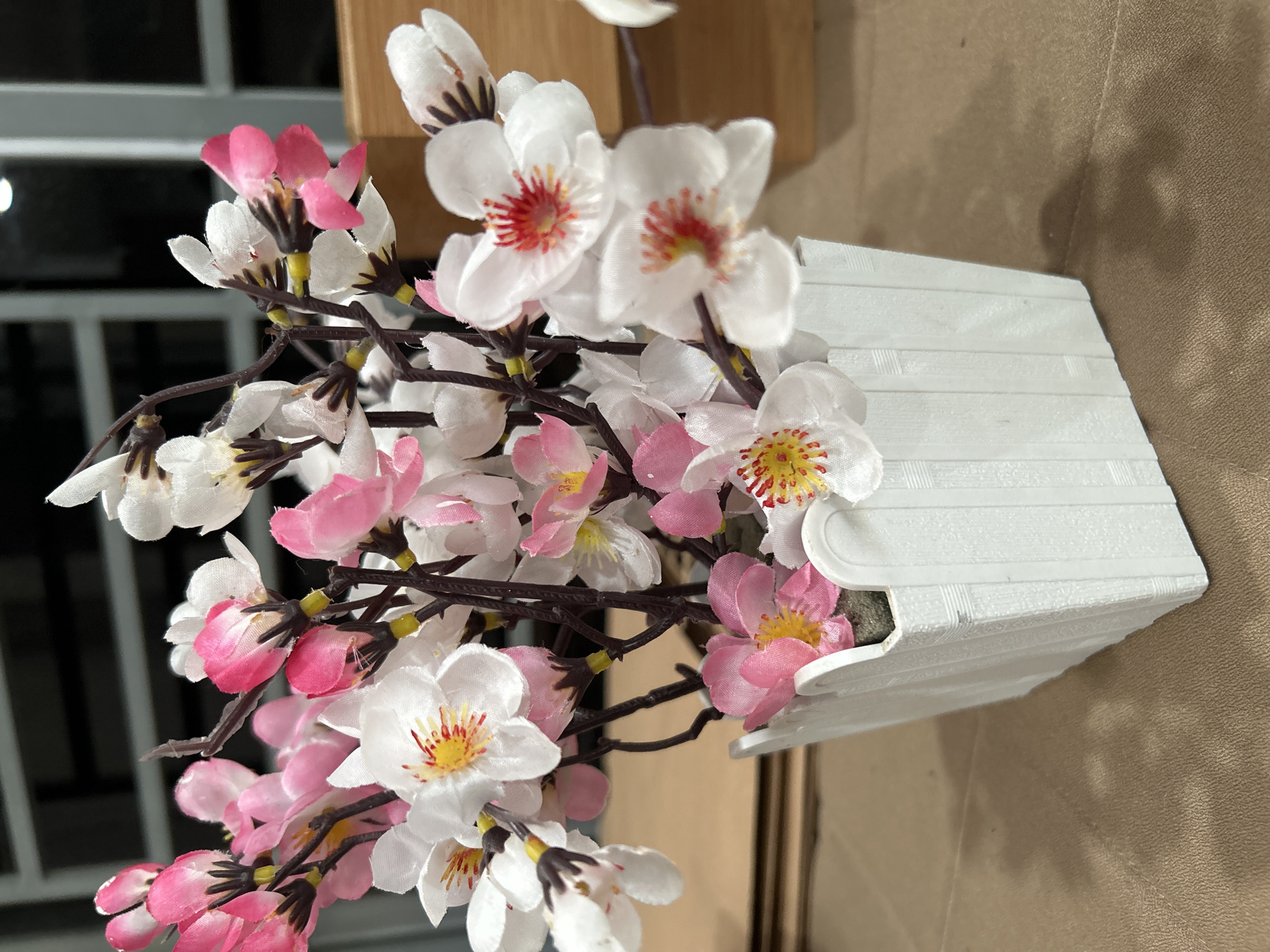 A close-up shot shows a bouquet of artificial pink and white cherry blossom flowers arranged in a small, rectangular white vase. The vase has a simple, textured design resembling horizontal planks. The flowers have brown stems and are positioned at various angles, creating a full and vibrant display. The background is a combination of light and dark areas, suggesting an indoor setting near a window or a dimly lit space. The location in Lhokseumawe, Indonesia, is not discernible from the image.