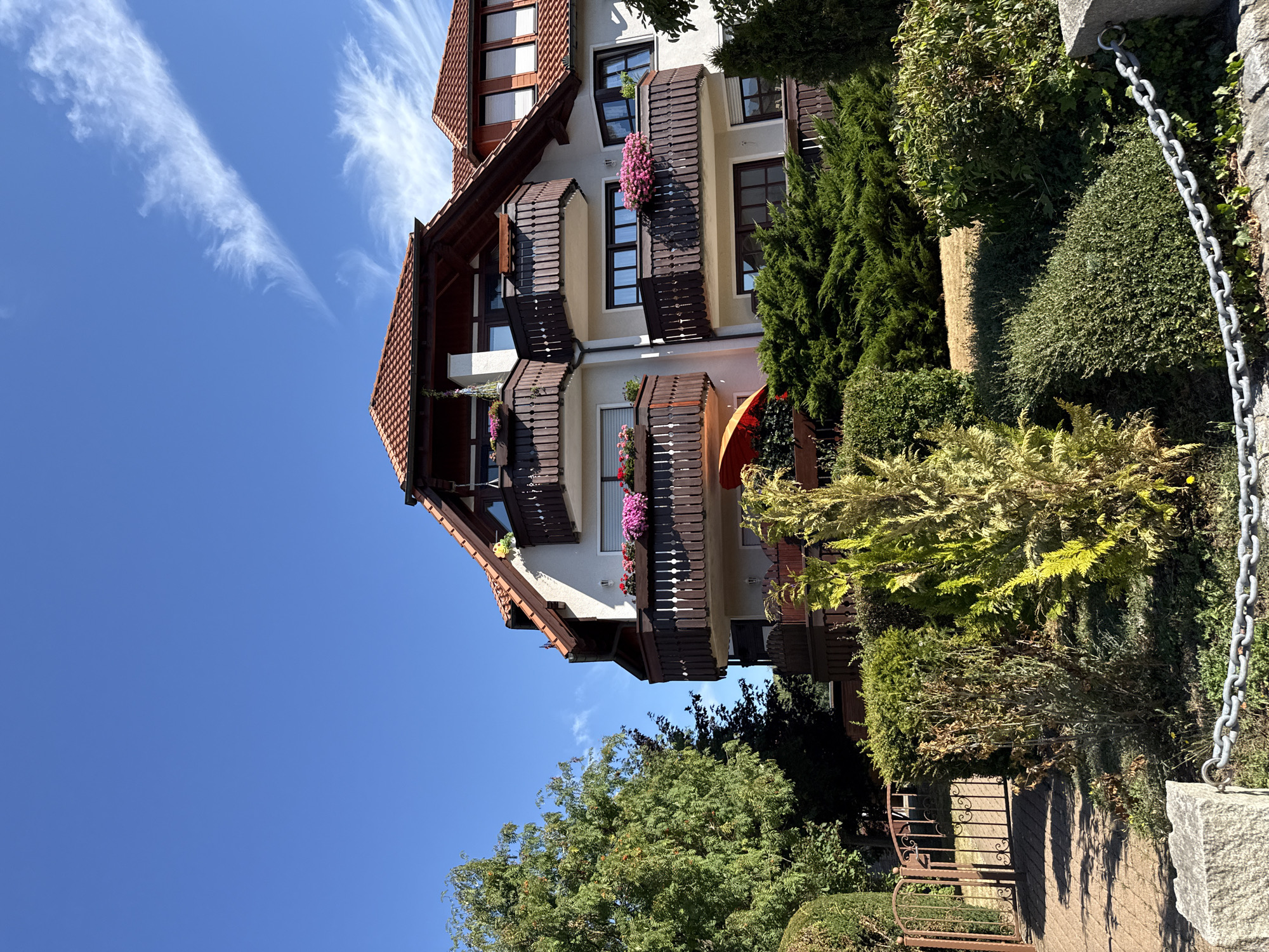 This image, taken in Bad Sachsa, Germany, depicts a multi-story residential building with a red tile roof and several balconies. The building's facade is white, and the balconies have dark wooden railings adorned with planters filled with pink flowers. In the foreground, there are various types of manicured shrubs and trees, including a tall, light green conifer and a smaller, dense green bush. To the left, an ornate, dark brown metal gate with decorative scrollwork is visible, leading to a paved walkway. A thick metal chain runs across the lower part of the image, attached to stone posts. The sky is a clear, bright blue with wispy white clouds. The scene appears to be a sunny day, likely in the summer or early autumn, given the foliage and the presence of an orange umbrella visible on one of the balconies. There are no people visible in the image.