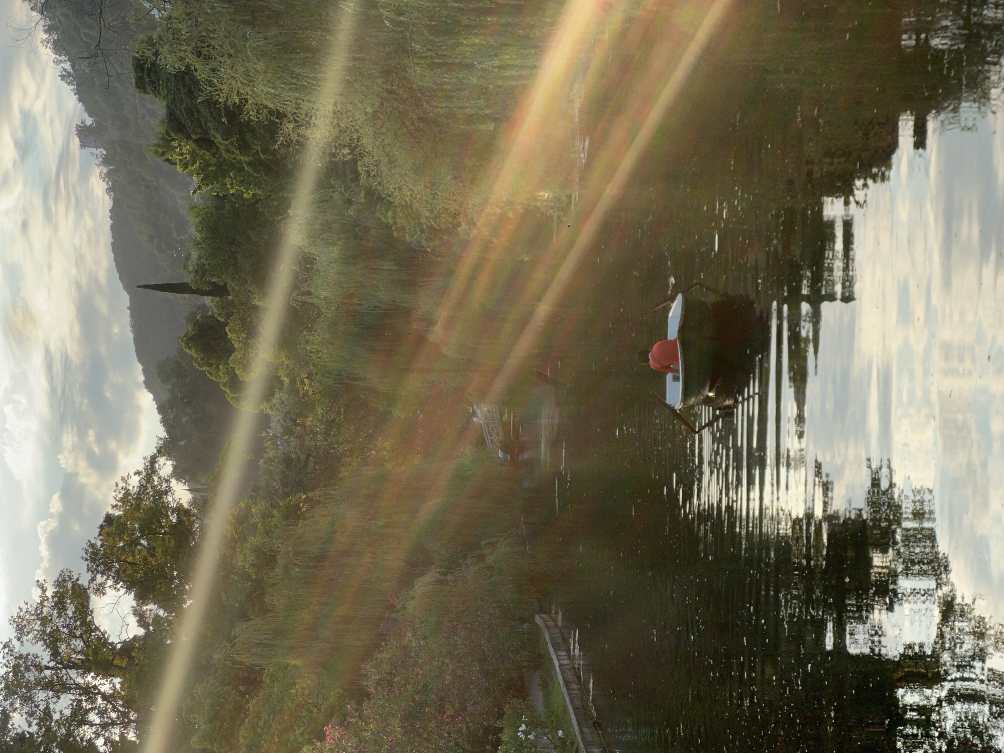 In the city of New Athos, Abkhazia, a lone individual wearing a red shirt rows a boat through a calm body of water. The scene is bathed in dramatic sunlight, with strong rays of light creating a lens flare effect across the image. The water reflects the surrounding lush greenery, a mix of trees and bushes that cover the gentle slopes of the hills in the background. A narrow stone embankment lines the left side of the waterway, with vegetation growing up to its edge. In the distance, a bridge crosses the water, and further back, the landscape rises into rolling hills covered in dense forest. The sky above is partly cloudy, with the sunlight filtering through the clouds.