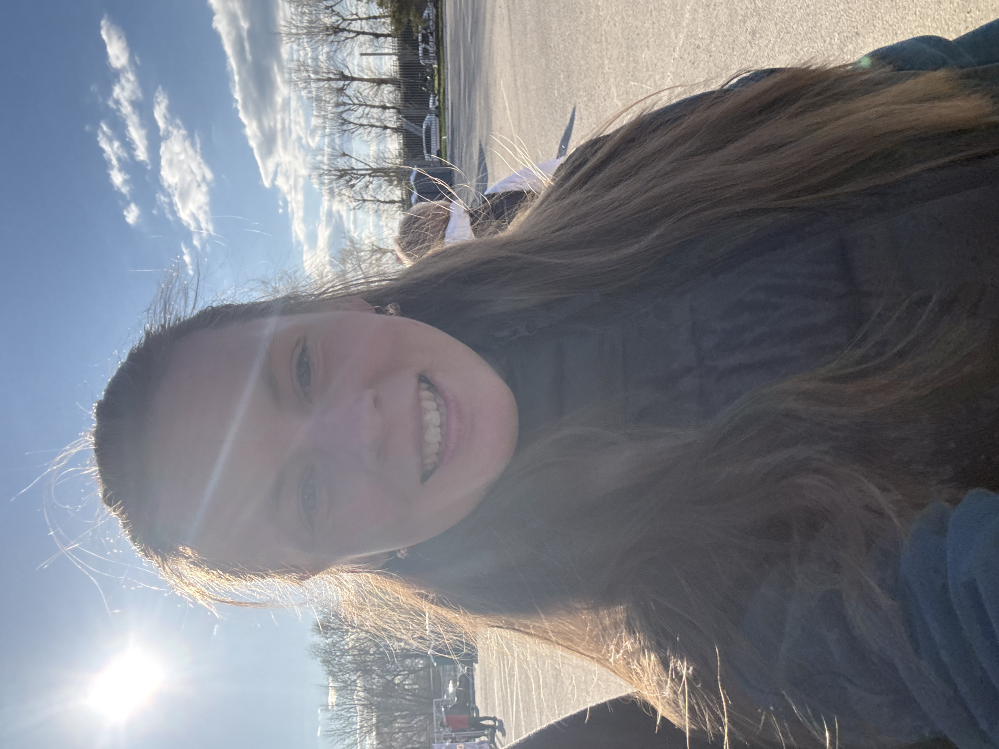 A young woman with long brown hair smiles at the camera. She is outdoors on a sunny day. The sky is blue with some white clouds. Bare trees are visible in the background. There is also a paved area and possibly some vehicles. The location is Kazan, Russia.
