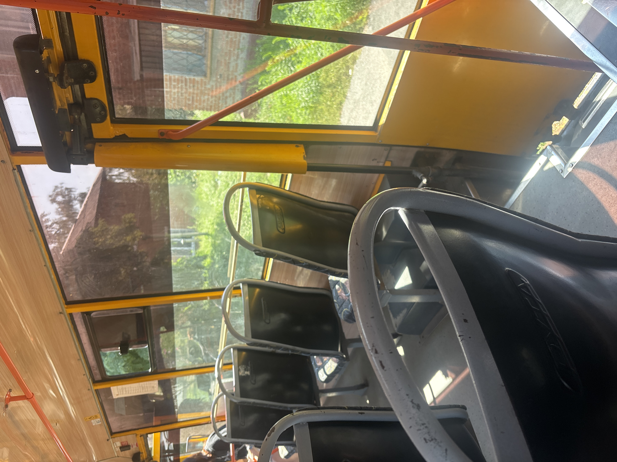 An interior shot captures the seating area of a public bus, likely in Kremenchuk, Ukraine, on a sunny day. The seats, upholstered in a patterned fabric, are visible alongside the bus's yellow interior frame and windows. Sunlight streams through the windows, illuminating the aisle and casting shadows. The external view shows buildings and greenery. The image focuses on the bus's interior, with a partial view of a person on the left.