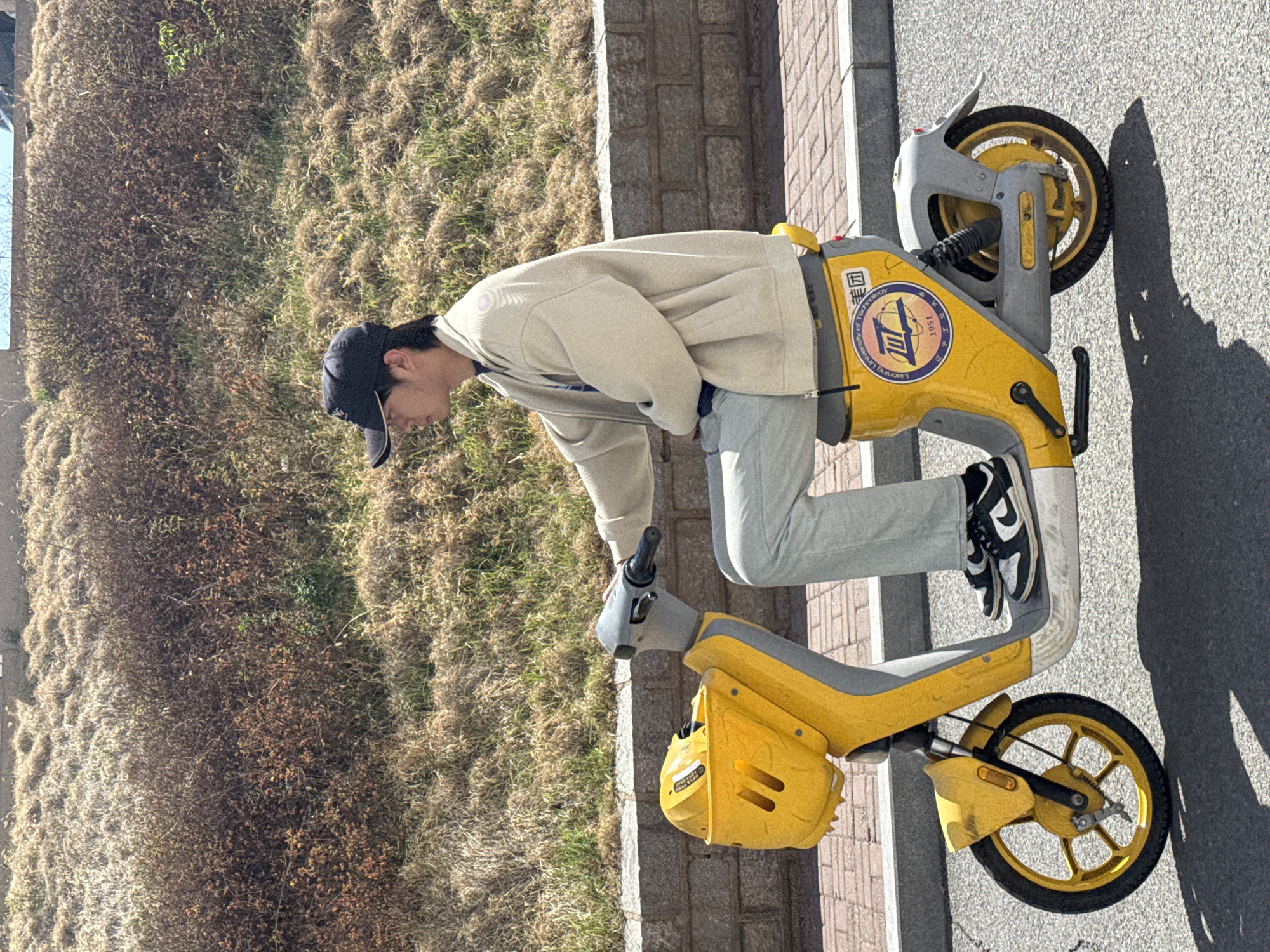 A young man wearing a black baseball cap, a beige jacket, and light grey pants is bending over a yellow scooter parked on a paved surface. The scooter has a "Liaoning University of Technology" logo on its side. The location appears to be an outdoor urban area, possibly in or near Zhongtun, China, based on the university logo on the scooter. To the side of the scooter, there is a patch of dry grass and a low concrete wall. The ground surface changes from pavement to brick near the scooter and grass.