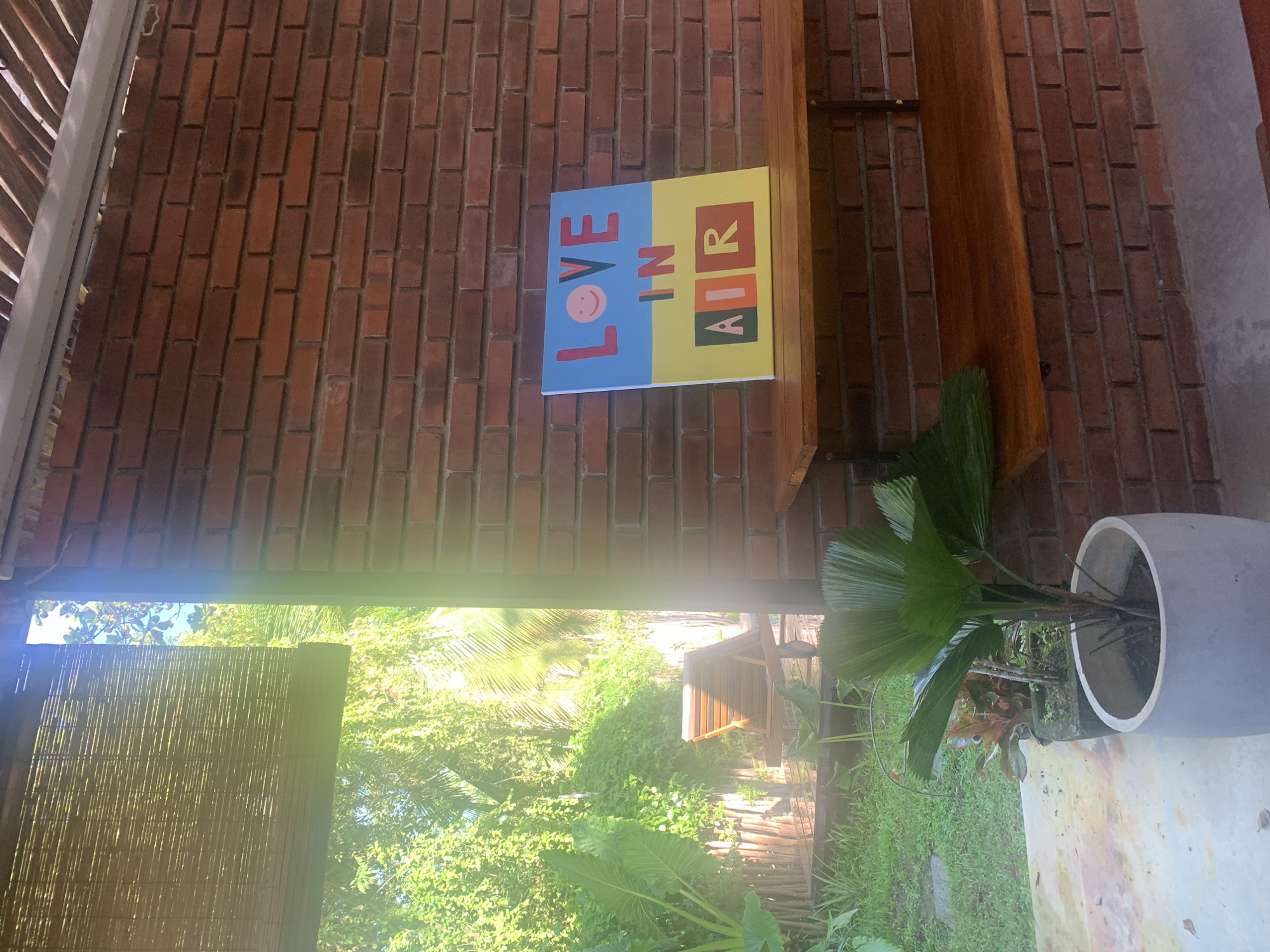 An outdoor scene in Kabalana, Sri Lanka, features a brick wall with a wooden shelf displaying a painting. The painting, predominantly blue and yellow, features the text "LOVE IN AIR" with a smiley face. To the left of the wall, lush green foliage and a sun lounger are visible through a bamboo blind and an open doorway. In the foreground, a potted plant with large green leaves sits on a concrete floor.
