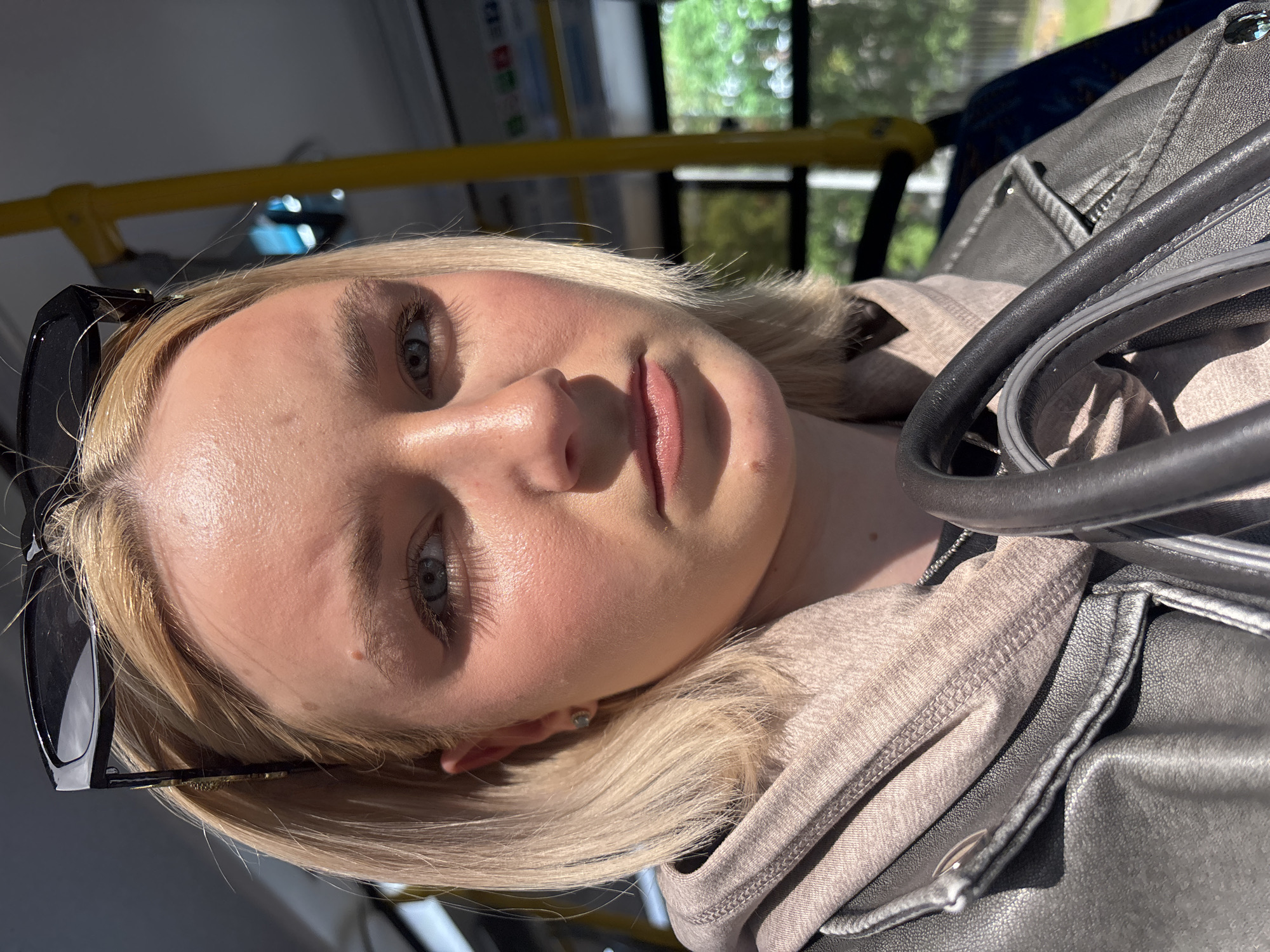 This is a portrait of a young woman with blonde hair, taken from a low angle, likely inside a public transportation vehicle given the yellow grab pole and interior visible in the background. The woman is wearing sunglasses on her head, a light-colored hooded garment, and a dark leather jacket. Her face is partially illuminated by sunlight, casting shadows and highlighting her features, including her blue eyes and pink lips. She appears to be looking at the camera, with a neutral or slightly curious expression. The overall lighting suggests it is daytime, possibly late morning or early afternoon, with bright sunlight filtering in from outside. The scene is indoors but with views of greenery through windows, indicating an urban or suburban setting. There is no visible text in the image.