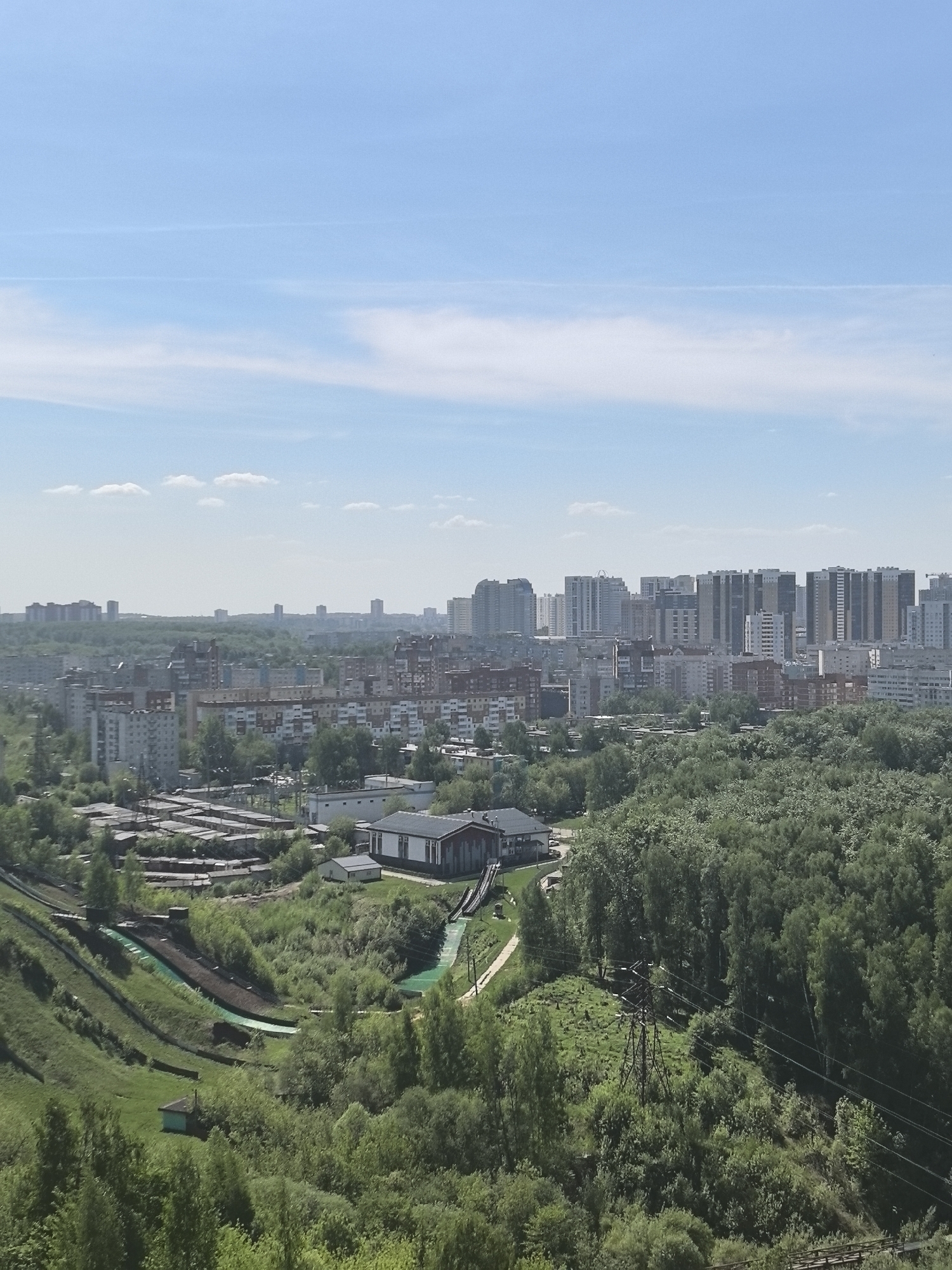 The image shows a cityscape with a dense urban area extending into the distance, transitioning to a foreground dominated by lush greenery. Tall residential buildings are visible on the horizon, interspersed with lower-rise buildings closer to the viewer. In the foreground, there's a ski jumping complex with artificial slopes in varying shades of green, indicating different sections of the ski jump facilities. There are support structures and maintenance buildings associated with the complex. Dense trees cover the majority of the foreground, with power lines running through the greenery. Above the city, a clear blue sky with a few scattered clouds is present. The image depicts the city of Perm, Russia, known for its urban landscapes and surrounding natural environment.