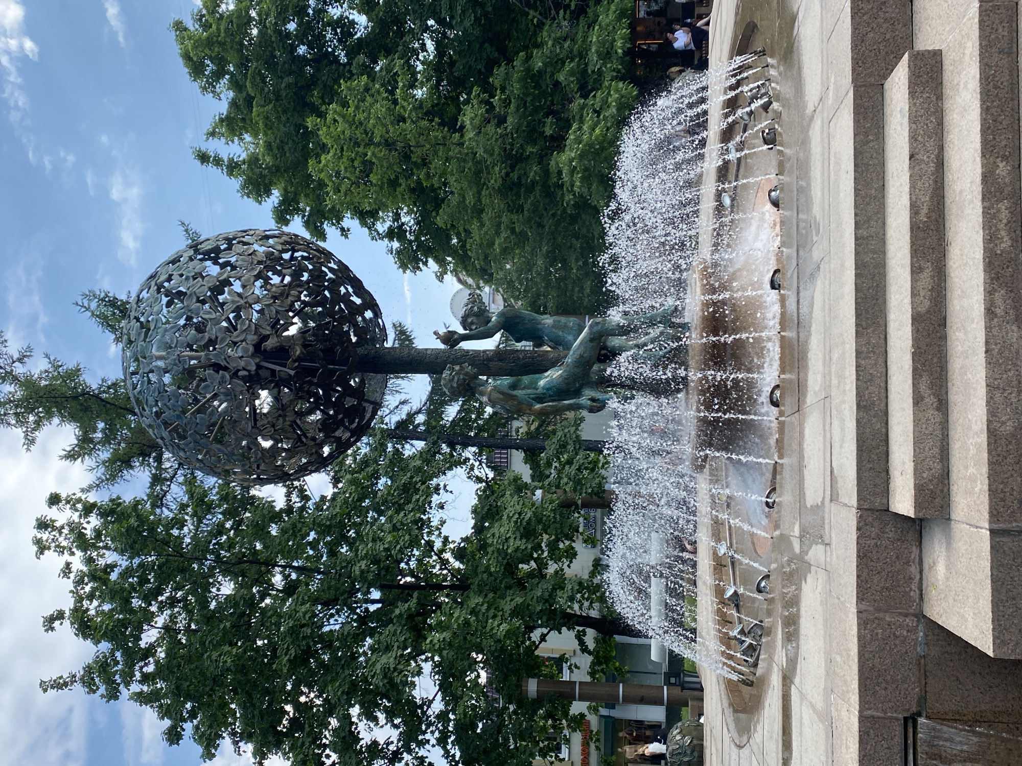 The image shows the "Triumph of Life" fountain in Moscow, Russia. The fountain features a bronze sculpture of two figures under a large metal sphere adorned with leaf patterns. Jets of water are spraying from the side of the fountain, which is built with gray stone. A lush green tree stands behind the fountain. The sky is a mix of blue and white clouds. People are visible in the background.