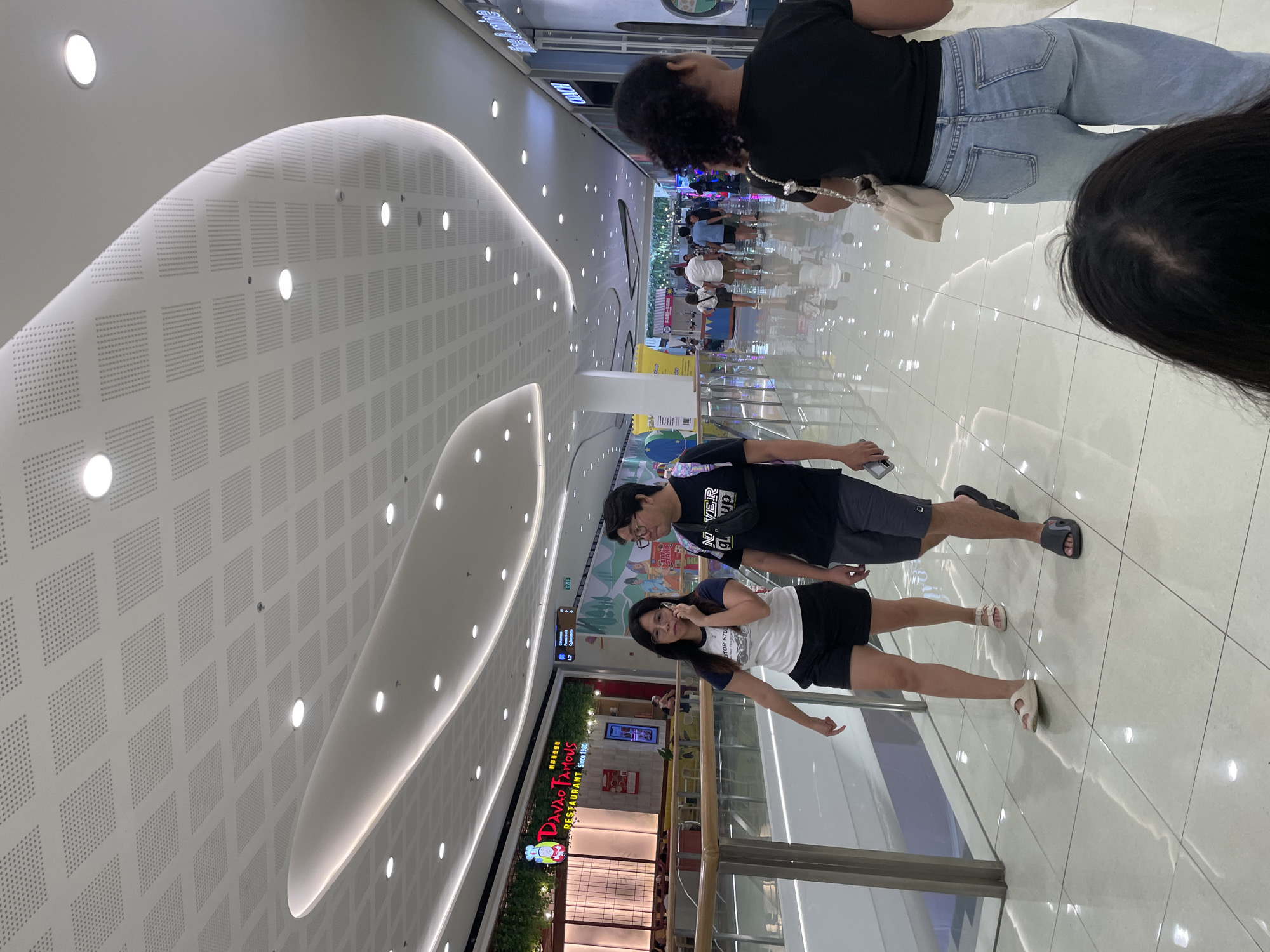 This image depicts an indoor scene within a modern shopping mall, identified by the "DAO TAO FAMOUS RESTAURANT Since 1990" sign and directional signage indicating "L2 Cinemas, Cyberzone," placing the location in Davao City, Philippines, likely SM Lanang Premier.
The environment features a white ceiling with a grid of perforated acoustic panels and numerous recessed circular LED lights. Curved, illuminated architectural elements with cove lighting enhance the ceiling design. The flooring consists of light-colored, polished tiles reflecting the overhead illumination. A metal and glass railing runs along the left side, delineating a walkway or upper level.
In the middle ground, two individuals walk towards the left of the frame. A woman on the left wears a white t-shirt with a dark graphic print, black shorts, and white flat sandals. She has dark hair, wears glasses, and holds a smartphone to her right ear, appearing to be on a call. Beside her, a man wears a dark t-shirt with a white graphic, dark grey shorts, and dark sandals. He wears glasses and a black fanny pack or sling bag across his chest, and holds a smartphone in his left hand.
On the right side of the foreground, the back of a person's head with dark, curly hair is partially visible, indicating the perspective of the shot. Further right, the upper body of this person is seen from behind, wearing a black short-sleeved t-shirt and light blue denim jeans. A light-colored shoulder bag with a silver chain strap is slung over their right shoulder.
The background reveals more of the mall interior with several other individuals walking. The "DAO TAO FAMOUS RESTAURANT" has a warm-toned facade with wood paneling, green artificial foliage above its sign, and interior lighting. Further down the right side, a "COACH" store sign is partially visible. The overall scene is brightly lit and indicates an active commercial setting.