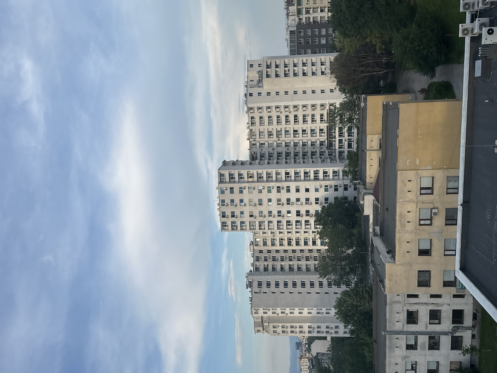 The image shows a cluster of apartment buildings in a city, likely Sofia, Bulgaria, under a cloudy sky. The main focus is on several tall, white, monolithic apartment blocks with numerous windows and external air conditioning units. In the foreground, there are lower, older buildings with worn facades, one painted yellow and another a muted gray. Trees are interspersed throughout the scene, with dense foliage surrounding some of the structures. The overall impression is of a dense urban residential area.