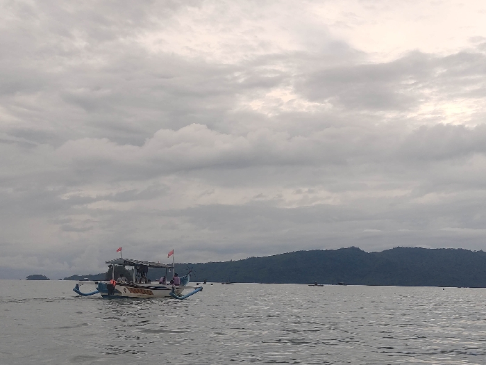 The image depicts a serene marine landscape in Watulimo, Indonesia, under a heavily overcast sky. A traditional Indonesian outrigger boat, likely a jukung, is the prominent subject in the mid-ground, positioned slightly left of center. This boat features a central covered structure with two small red flags flying from its roof. Several people are visible seated within the boat under the canopy, though their faces and specific activities are indistinct. Outrigger floats extend from both sides of the vessel, maintaining its stability on the water. Faint white text is visible on the hull of the boat, but it is illegible.
The water surface is calm with gentle ripples, reflecting the subdued light from the sky. In the background, a long, verdant landmass composed of rolling hills or mountains stretches across the horizon. Several smaller boats are visible as dark silhouettes near this distant coastline. The sky is entirely covered with thick, grey clouds, indicating a cloudy day, likely mid-morning or afternoon, as there is no harsh sunlight or strong shadows. The overall atmosphere conveyed is one of tranquility and peace, characteristic of a quiet moment on the water in a tropical region.
