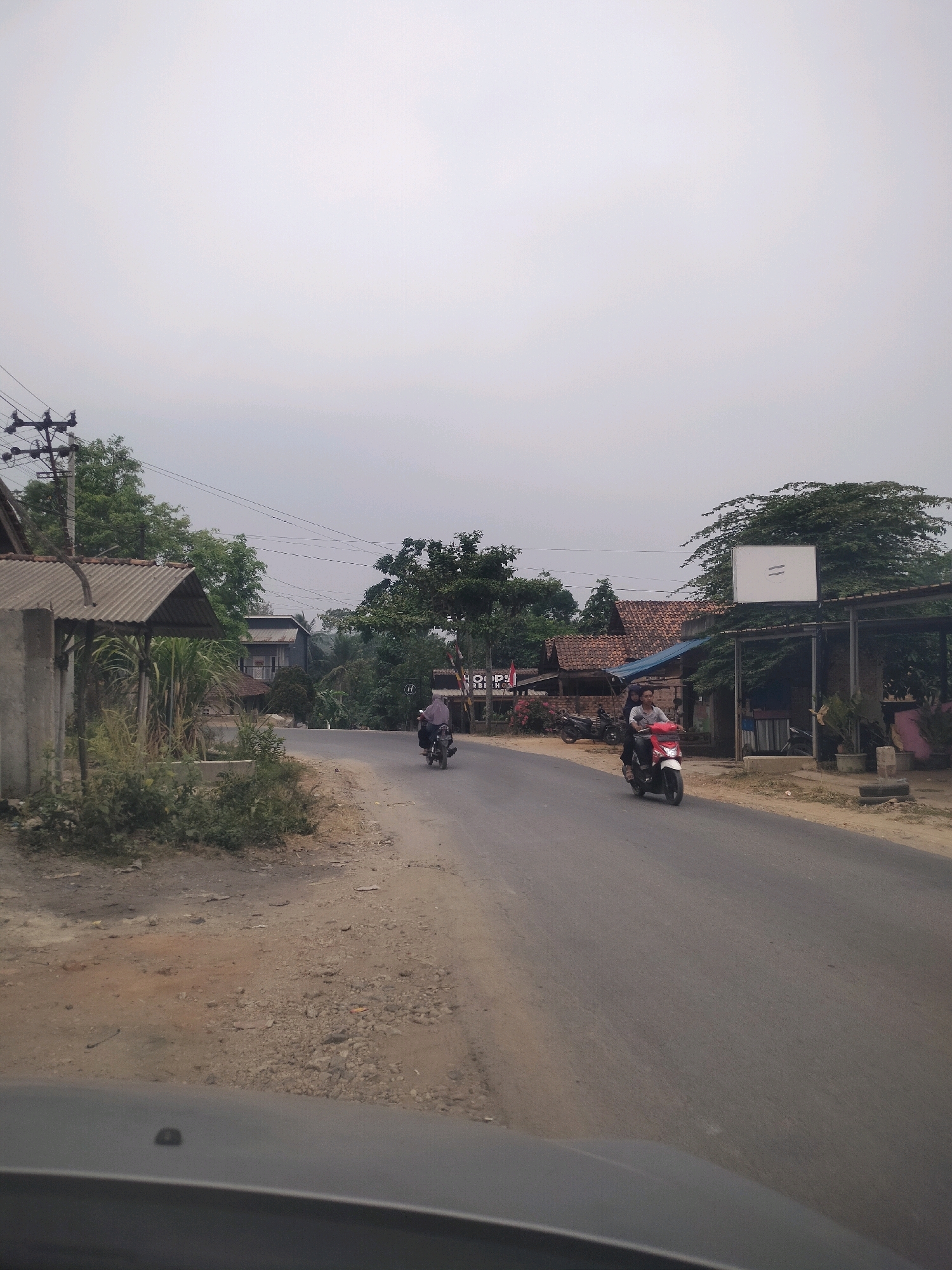The image depicts a street scene in Wayakroy, Indonesia. Two individuals are riding a red scooter on the right side of the asphalt road, moving away from the viewer. The scooter has a white front fender and white sides. The driver of the scooter is wearing a white shirt and dark pants, and the passenger is wearing a blue shirt and dark pants. On the left side of the road, a motorcycle with a single rider is also moving away from the viewer. The rider is wearing a light-colored shirt and dark pants.
The environment includes several buildings with traditional Indonesian architecture, featuring terracotta tile roofs. Trees and lush vegetation are visible throughout the scene. Utility poles with electrical wires are present on the left side of the road. A sign with a white rectangular surface and two horizontal lines in black is mounted on a pole above a covered area on the right side of the road. The road surface appears to be asphalt with some gravel and dirt on the shoulder. The sky is overcast, suggesting a cloudy day. The foreground shows a portion of a gray vehicle's dashboard, indicating the image was taken from inside a car.