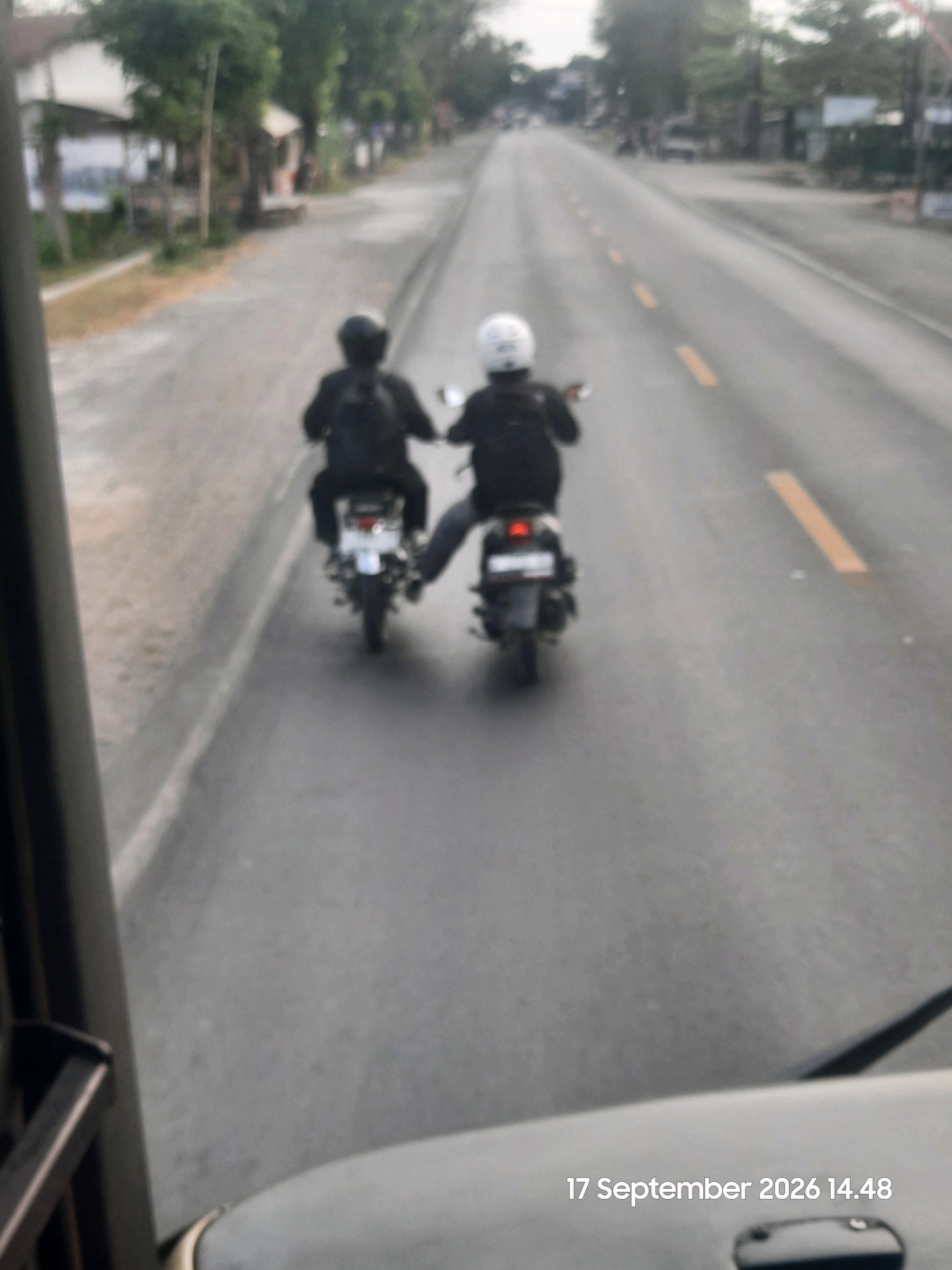 Two individuals are riding motorcycles side-by-side on a paved road. The person on the left is wearing a black helmet and a black backpack, and is riding a motorcycle. The person on the right is wearing a white helmet and is also riding a motorcycle. Both individuals are facing away from the camera. The road is lined with yellow dashed lines. On the left side of the road, there are buildings and trees. The scene appears to be in Sukokerto, Indonesia. The bottom right of the image shows a timestamp: "17 September 2026 14.48".