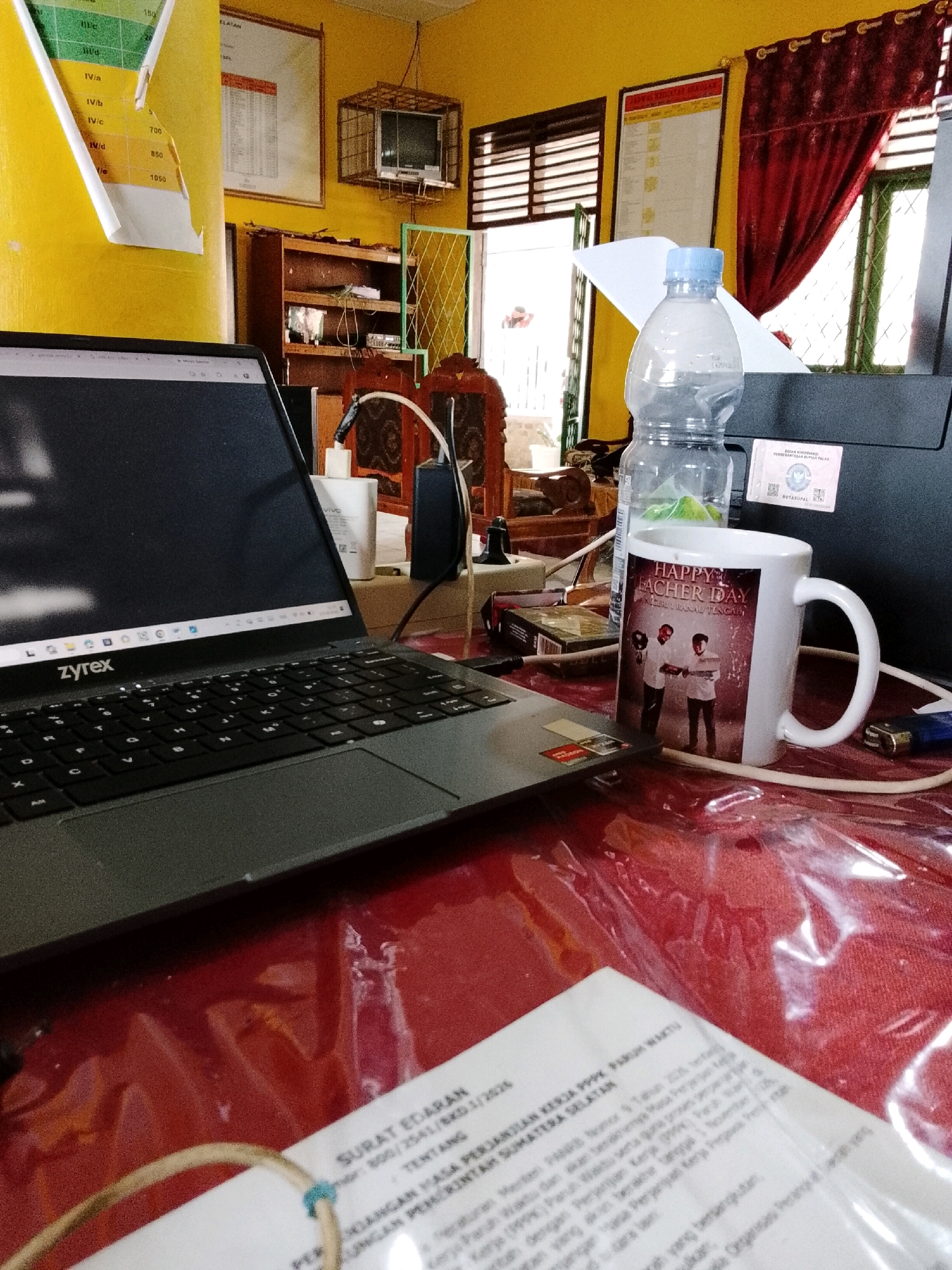 The image depicts a cluttered desk in what appears to be an office or study room. A laptop with the "zyrex" logo is open in the foreground, displaying a web browser. To the right of the laptop, there is a white mug with a "HAPPY FATHER DAY" design featuring two men. Behind the mug, a clear plastic water bottle is visible, along with a black folder and a sticker that reads "BADAN KOORDINASI PERESEPSI PEMERINTAH". A lighter is also present on the desk. In the background, there are shelves with various items, a framed document on the wall, dark red curtains, and a window with horizontal blinds. A piece of paper with Indonesian text, possibly a legal document, is partially visible in the lower right corner. The overall impression is one of a workspace in use, with personal items interspersed with work-related objects. The specific location of Jepara, Indonesia is not directly indicated by visual cues within the image.