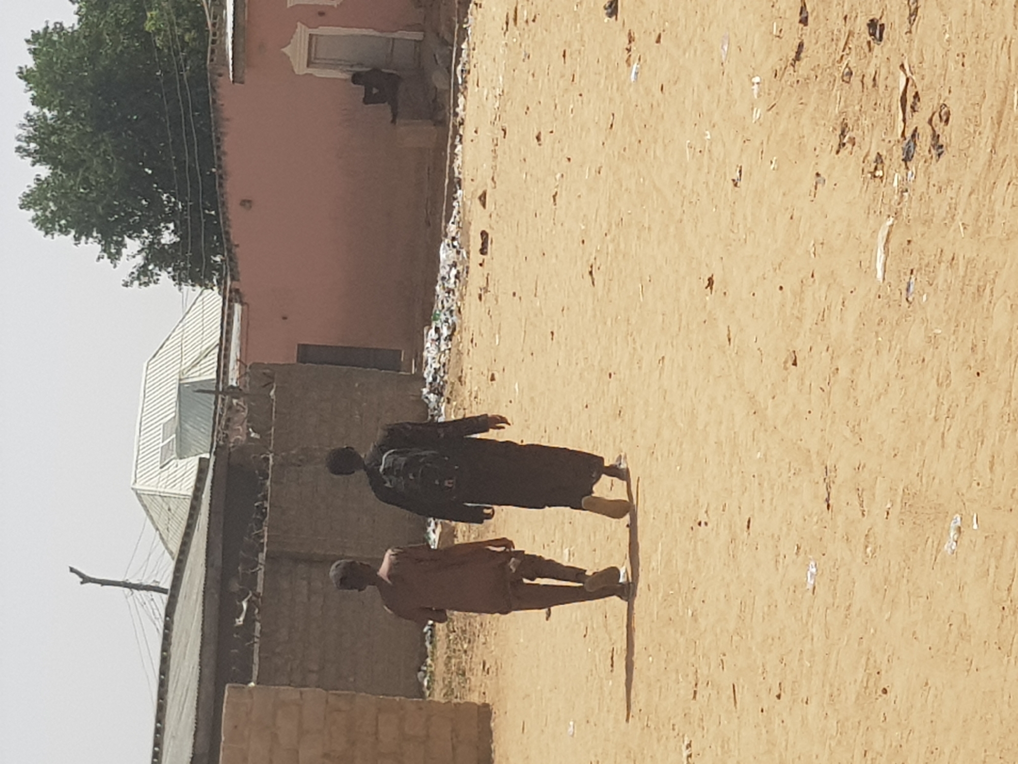 Two people are walking in a sandy area in Dabawa, Nigeria. One person is wearing a long, dark garment and carrying a backpack. The other person is wearing lighter-colored clothing. A building is to the left. A pink building is in the background. Debris is visible near the pink building. Another person is seated in the doorway of the pink building. A tree is near the pink building. Power lines extend from a pole to the building to the left.
