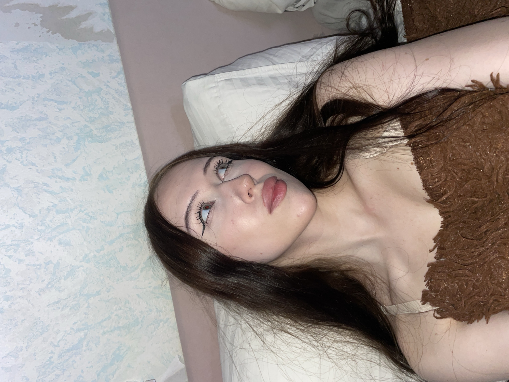 The media file is a close-up, low-angle photograph of a young woman lying on a bed. Her head is tilted back, and she is looking upwards. She has long, dark brown hair that is spread out around her. She is wearing a light-colored top, partially covered by a fluffy brown blanket. Her makeup includes dramatic eyeliner and mascara. The background consists of a textured wallpaper with a light blue and white pattern on the left, and a pinkish-beige wall on the right, with a white pillow supporting her head. The lighting appears to be artificial and soft, suggesting an indoor setting, possibly a bedroom. The overall mood is relaxed and perhaps contemplative. No text is visible in the image. The location context is Votkinsk, Russia.