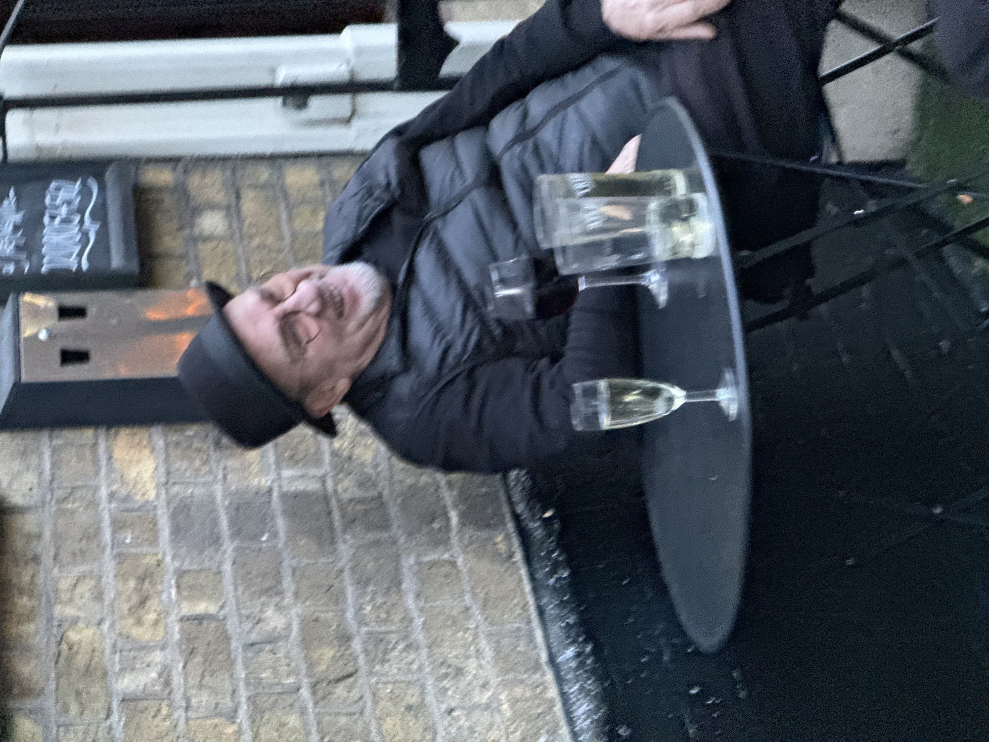A man is seated at a table with several glasses of liquid, likely champagne or sparkling wine. He is wearing a dark puffer jacket and a black hat. The table is round and black, positioned outdoors. The background shows a brick wall and a sign with writing on it. The location is potentially London, United Kingdom.