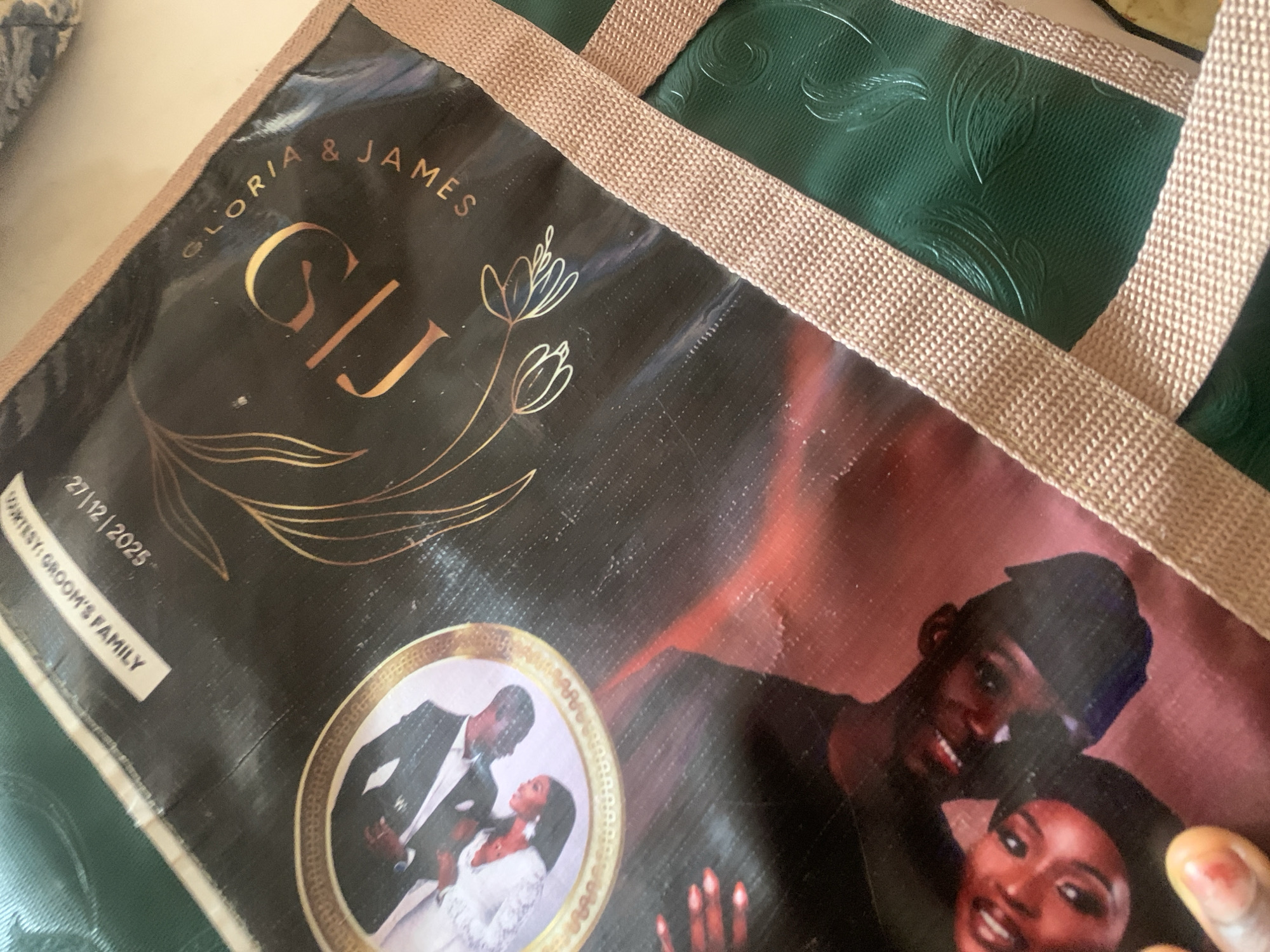 The image depicts a close-up view of a black fabric item, possibly a bag or a banner, adorned with a wedding design. Prominently displayed are the initials "SJ" in a large, ornate golden font, with "GLORIA & JAMES" written in smaller golden letters encircling them. A floral motif, also in gold, is visible to the left of the initials. A circular framed photograph of a wedding couple in formal attire is situated on the left side of the fabric. Below this, a partial image of a couple's faces is visible, with one face partially obscured by a black mask. Text at the top of the fabric reads "COURTESY: GROOM'S FAMILY" and "27/12/2025". The fabric also features a textured green panel with embossed leaf designs, bordered by a tan woven ribbon. The image quality is somewhat obscured by a plastic-like sheen on the fabric, and a portion of a hand with painted fingernails is visible in the bottom left corner. The setting is not identifiable as being in Jalingo, Nigeria, or any specific city.