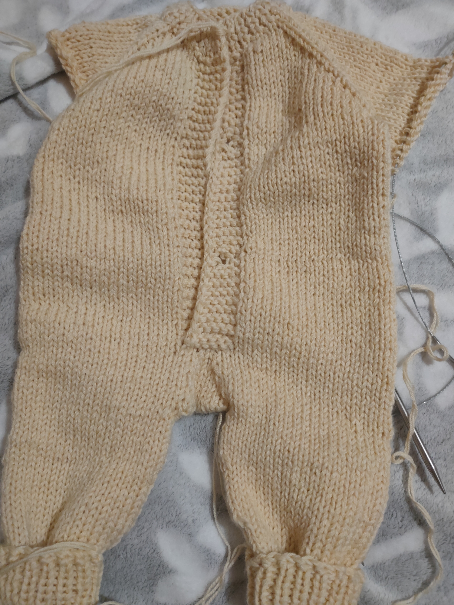 The image shows a cream-colored knitted baby romper laid flat on a gray and white patterned fabric. The romper is a one-piece garment with short sleeves, a button-up front, and long legs. The knitting appears to be done in a stockinette stitch with a ribbed cuff at the bottom of each leg. Several loose strands of yarn and a pair of knitting needles are visible next to the romper. The lighting is natural, casting soft shadows. There are no people or animals present in the image, and the setting is a flat surface, likely a table or floor. The image is a close-up view, focusing entirely on the knitted garment and knitting supplies. There is no information available to determine the location in the city of Oktyabrsky, Russia.