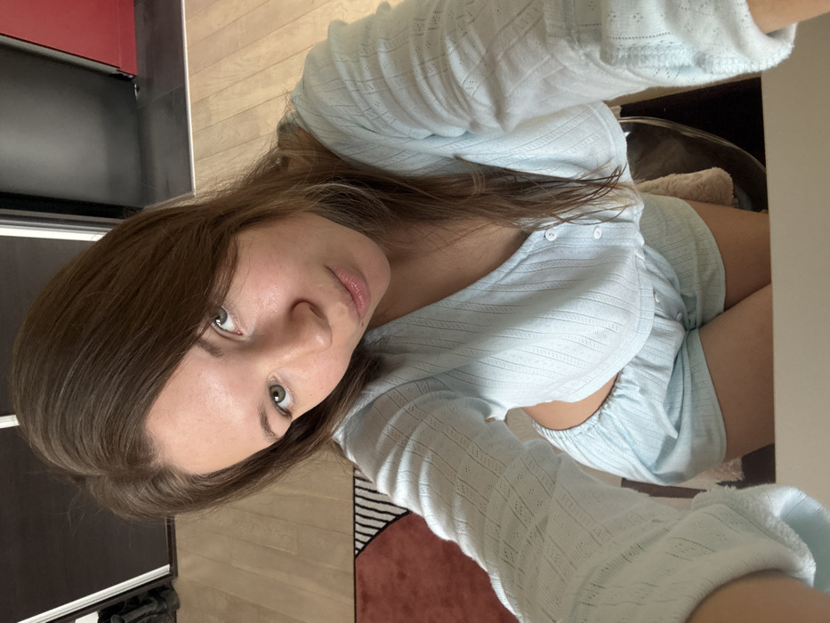 An indoor, high-angle, selfie-style photograph displays a light-skinned female individual with medium-length brown hair and green eyes. She is wearing a light blue, textured, cropped, long-sleeved top with a V-neck and white buttons, paired with matching light blue shorts. The individual gazes directly at the camera with a subtle pout. The immediate background includes light wooden flooring, a partial view of a rug featuring terracotta, black, and white geometric patterns, and dark-colored sliding doors. A portion of a dark wall with a red accent is visible on the far right. A white surface, likely a desk or table, is present in the foreground. A dark chair with a light-colored, soft covering is partially visible behind the individual's lower body. The specific location within the city of Vozdovac Urban Municipality, Serbia, cannot be determined from the image.