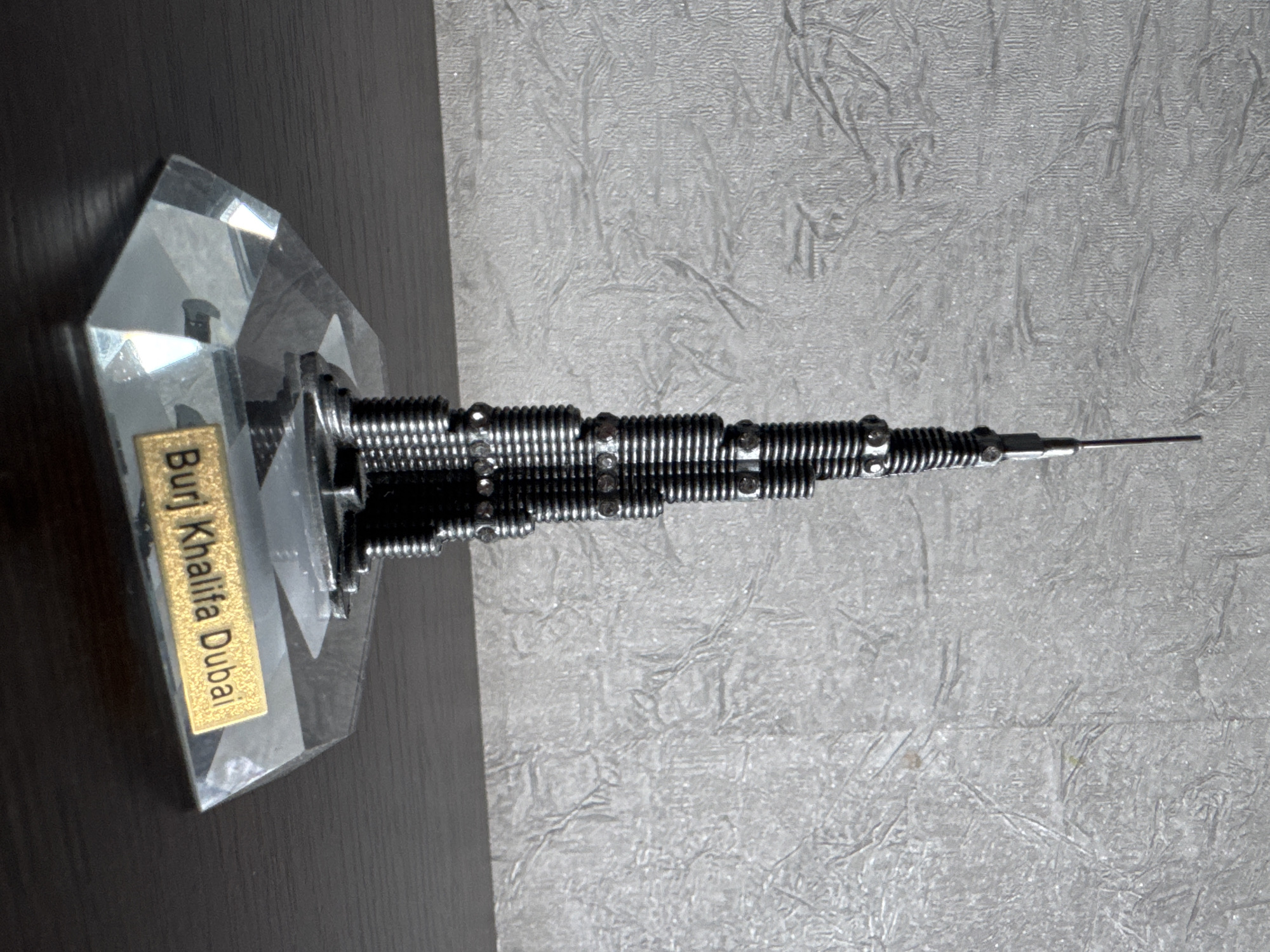 A miniature replica of the Burj Khalifa, a skyscraper, stands on a faceted clear acrylic base with a golden plaque that reads "Burj Khalifa Dubai". The replica is made of dark metal, possibly steel, and is adorned with small clear stones, resembling rhinestones, at intervals. The background is a textured grey wall. The replica is positioned on a dark brown wooden surface, likely a table. The image is a medium shot, focusing on the replica and its immediate surroundings. The lighting appears to be natural or soft artificial light, casting subtle shadows. There is no indication of the location being in Яромінскі сельскі Савет, Belarus.