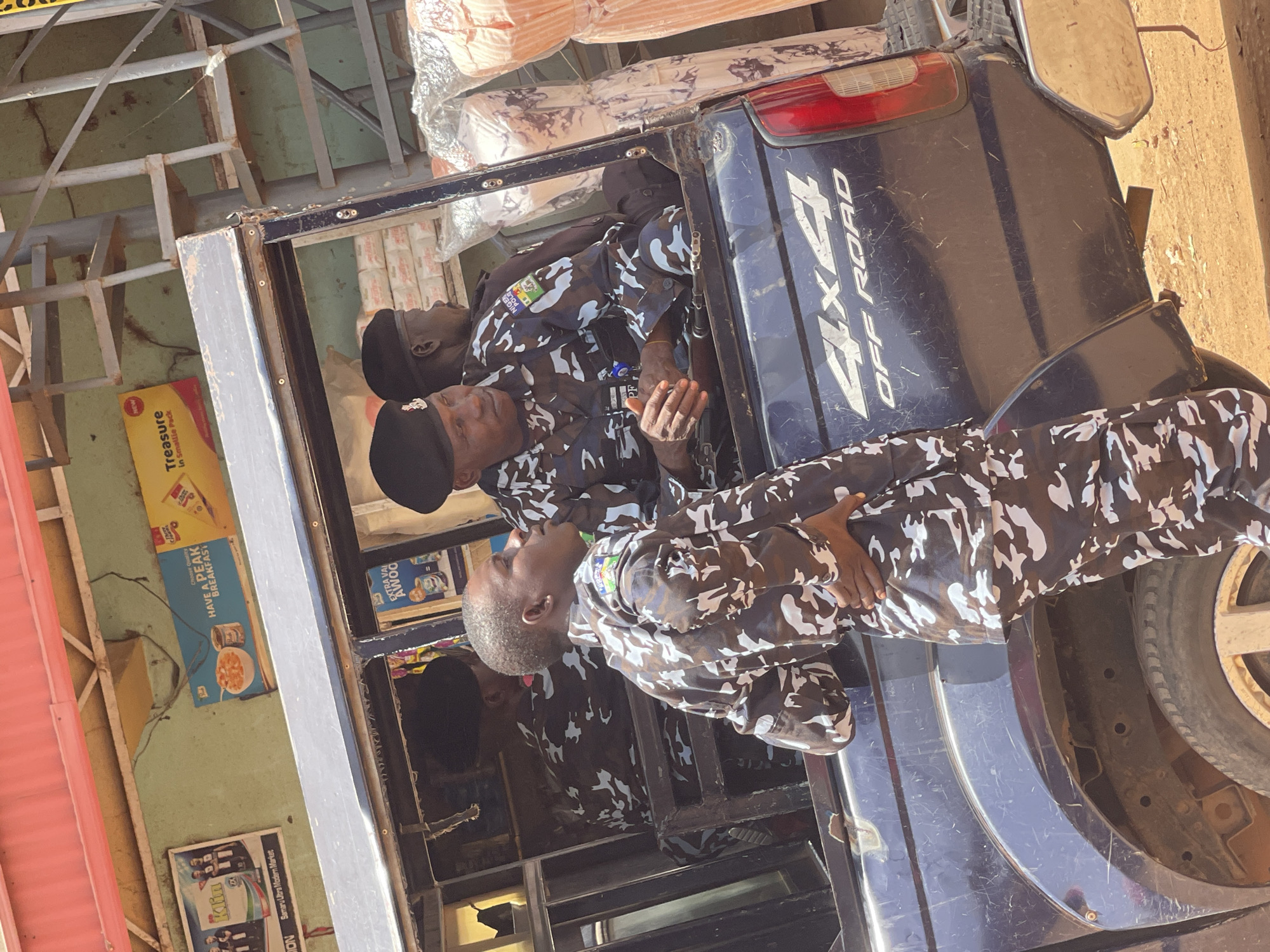 The image shows three men in camouflage uniforms seated in the back of a dark blue truck with "4x4 OFF ROAD" in white letters on the side. The men appear to be Nigerian police officers due to the patch on their uniforms. The scene appears to be outdoors with a building visible in the background, showcasing signs for "Treasure in Smallie Pack", "HAVE A PEAK BREAKFAST", and "Samaru Ultra Modern Market", indicating the location is in Samaru, Nigeria. The truck also contains what appears to be goods or luggage in the back.