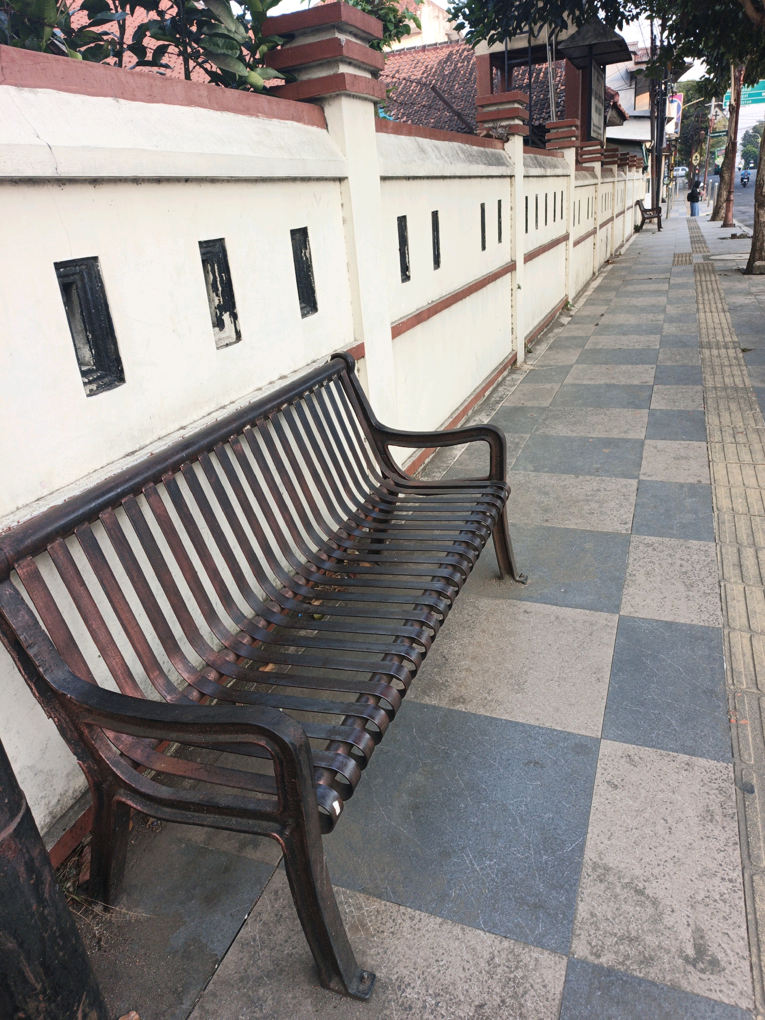 An eye-level, medium-wide shot depicts a park bench in a city sidewalk. The bench is dark brown metal with slatted seating and back. It is positioned in front of a cream-colored wall with a reddish-brown trim. The wall has several rectangular openings. The sidewalk is paved with large square tiles in a checkerboard pattern of gray and beige. To the right of the tiled sidewalk is a narrower section of textured, yellow pavement. Trees line the right side of the sidewalk, casting shadows. In the background, there are buildings with tiled roofs and other elements of a city street, including a traffic light and a few people walking further down the sidewalk. The lighting suggests it is daytime.