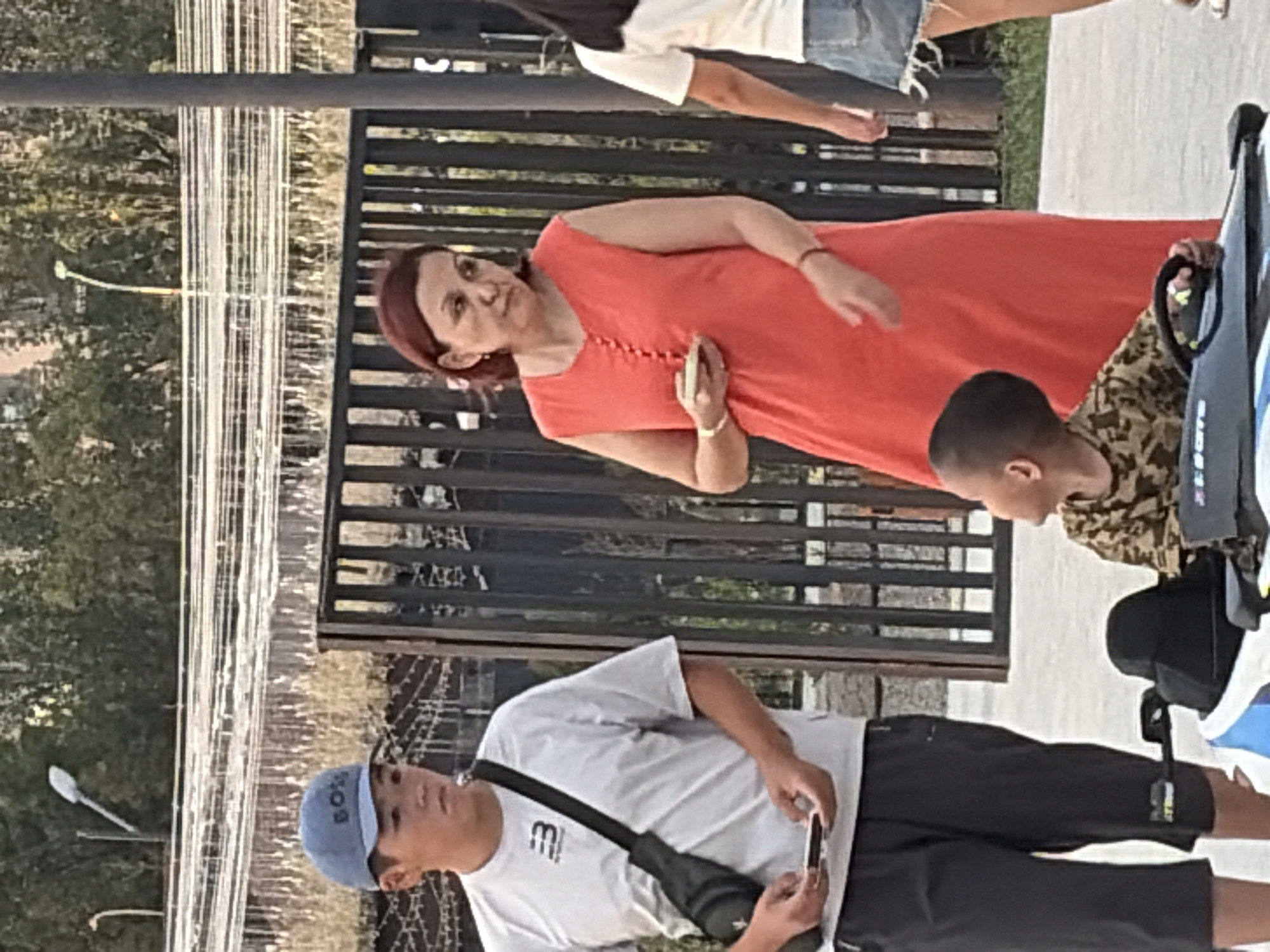 In the city of Unknown, Unknown, a young male wearing a grey t-shirt, black cap, and black pants stands on a paved surface. He carries a black bag across his chest and holds a white object in his left hand. To his right, a woman in a bright orange dress stands holding a small white object in her hand, with a thin red bracelet on her left wrist. In the foreground, a young child sits in a toy car, wearing a camouflage shirt and looking upwards. The background features a dark metal fence and trees. A street lamp is visible in the upper left portion of the image.