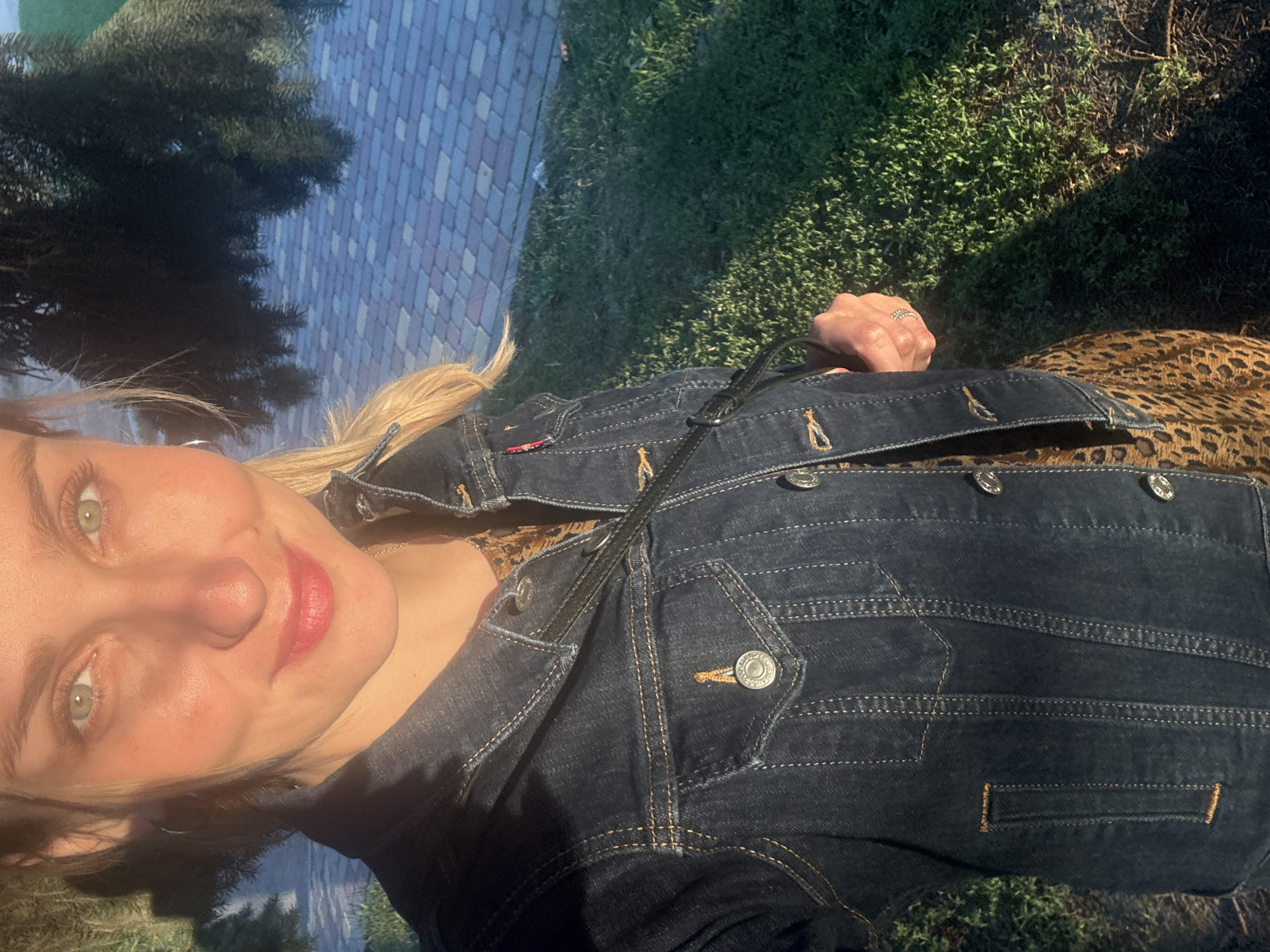 A woman with blonde hair and green eyes, wearing a dark denim jacket over a leopard-print dress, stands outside on a sunny day. She is holding a black purse with a strap that goes across her body. The background shows a paved walkway and green grass. The lighting suggests it is late afternoon or early morning.